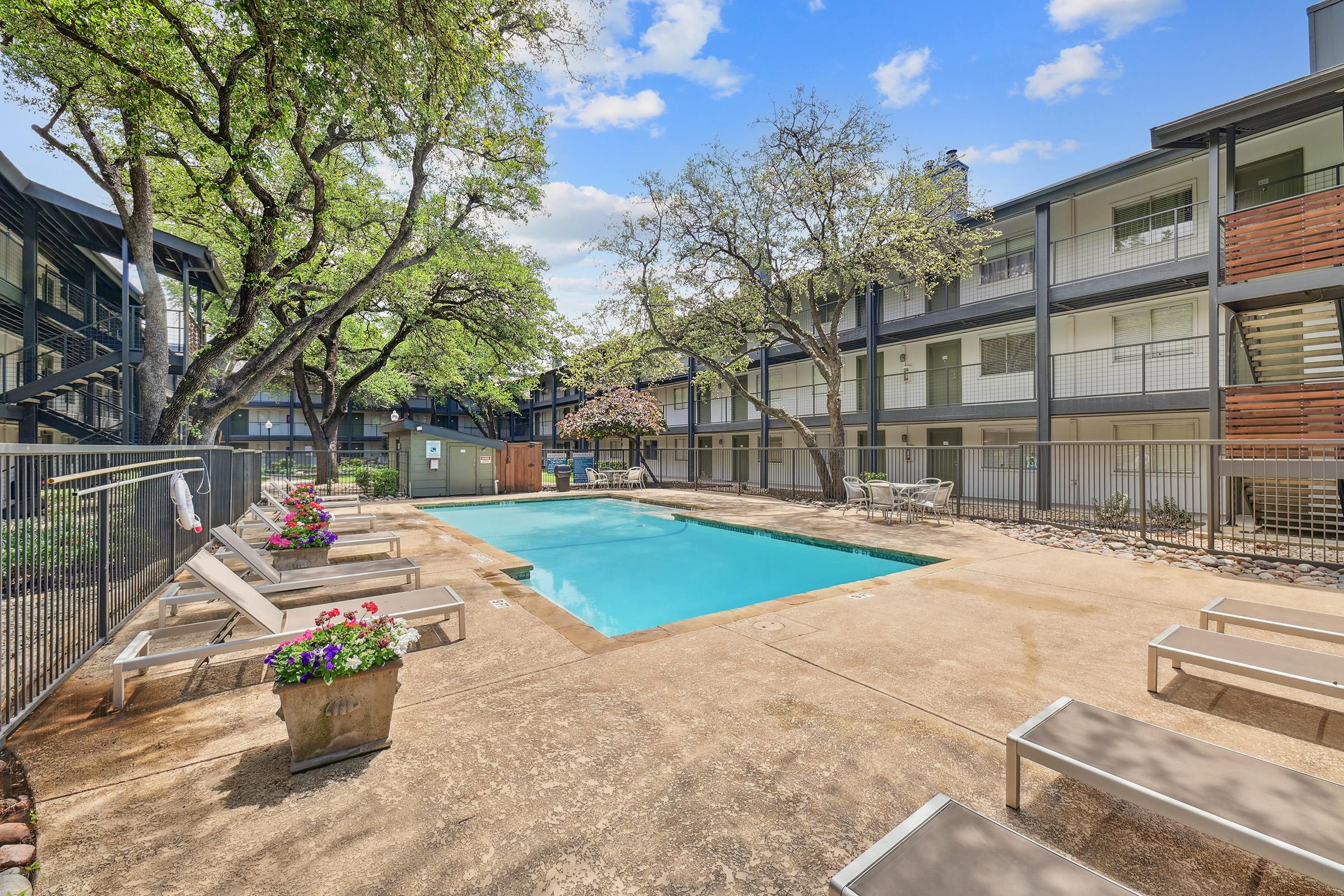 A swimming pool surrounded by lounge chairs and flower pots, set in a courtyard with lush green trees. Nearby buildings feature balconies and large windows, creating a serene and inviting outdoor space. Bright blue sky with a few fluffy clouds adds to the tranquil atmosphere.