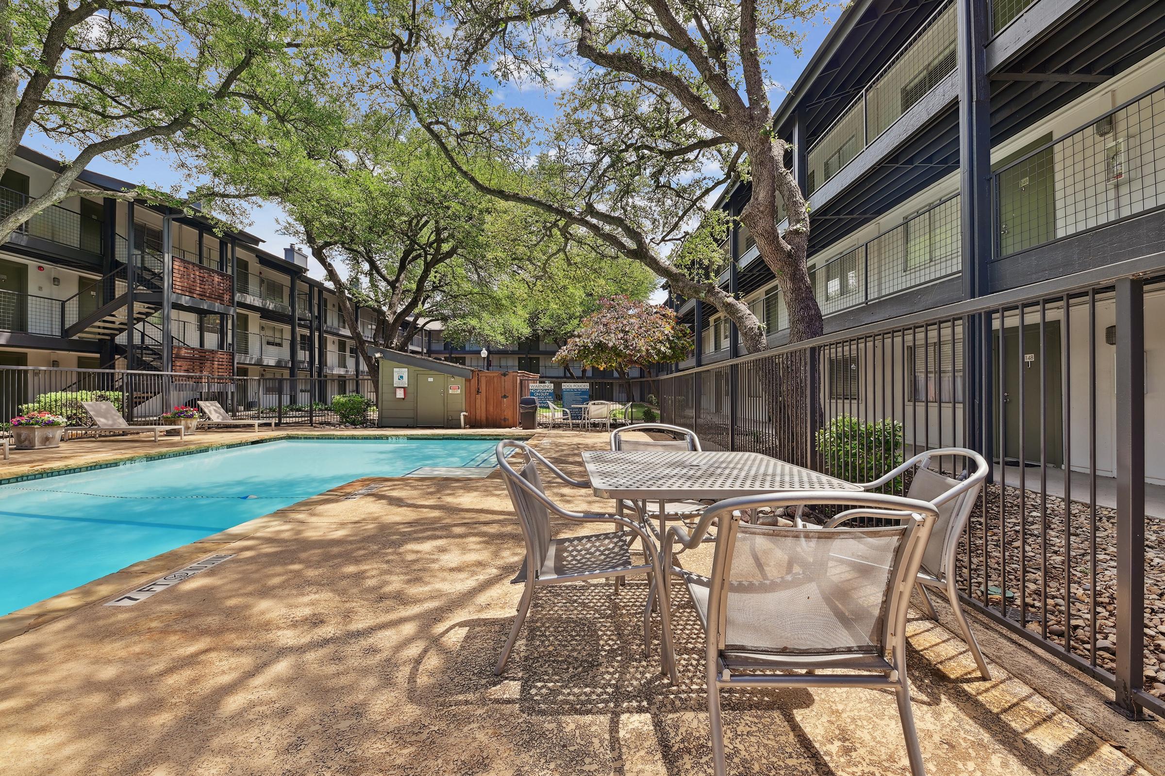A swimming pool surrounded by trees and an apartment complex. There are several outdoor tables and chairs near the pool, with steps leading to the upper levels of the apartments. The setting is bright and inviting, ideal for relaxation.