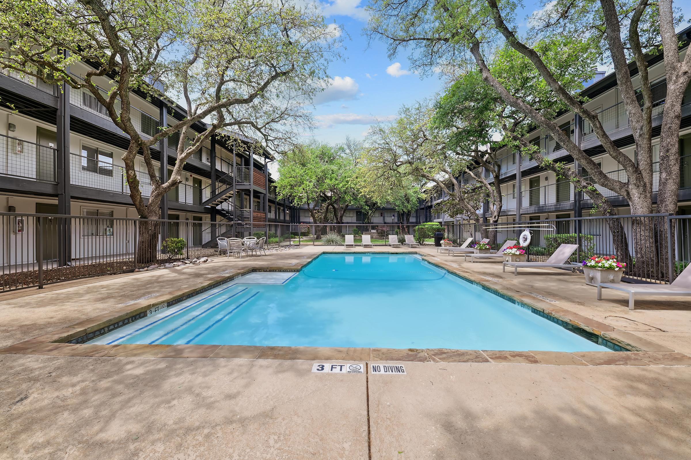A tranquil pool area surrounded by greenery, with well-maintained apartments in the background. The pool features a shallow end marked "3 FT" and has lounge chairs and flower pots nearby. Trees provide shade, creating a relaxing outdoor space. Clear blue sky enhances the inviting atmosphere.