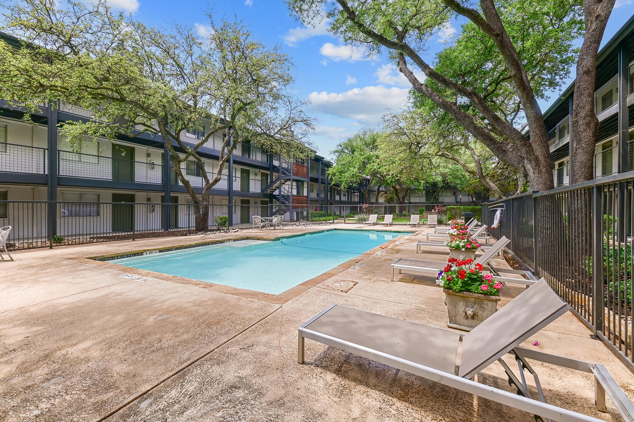 A tranquil courtyard featuring a swimming pool surrounded by lounge chairs, with potted flowers adding color. The area is shaded by trees and flanked by two-story buildings, creating a serene and inviting atmosphere for relaxation.