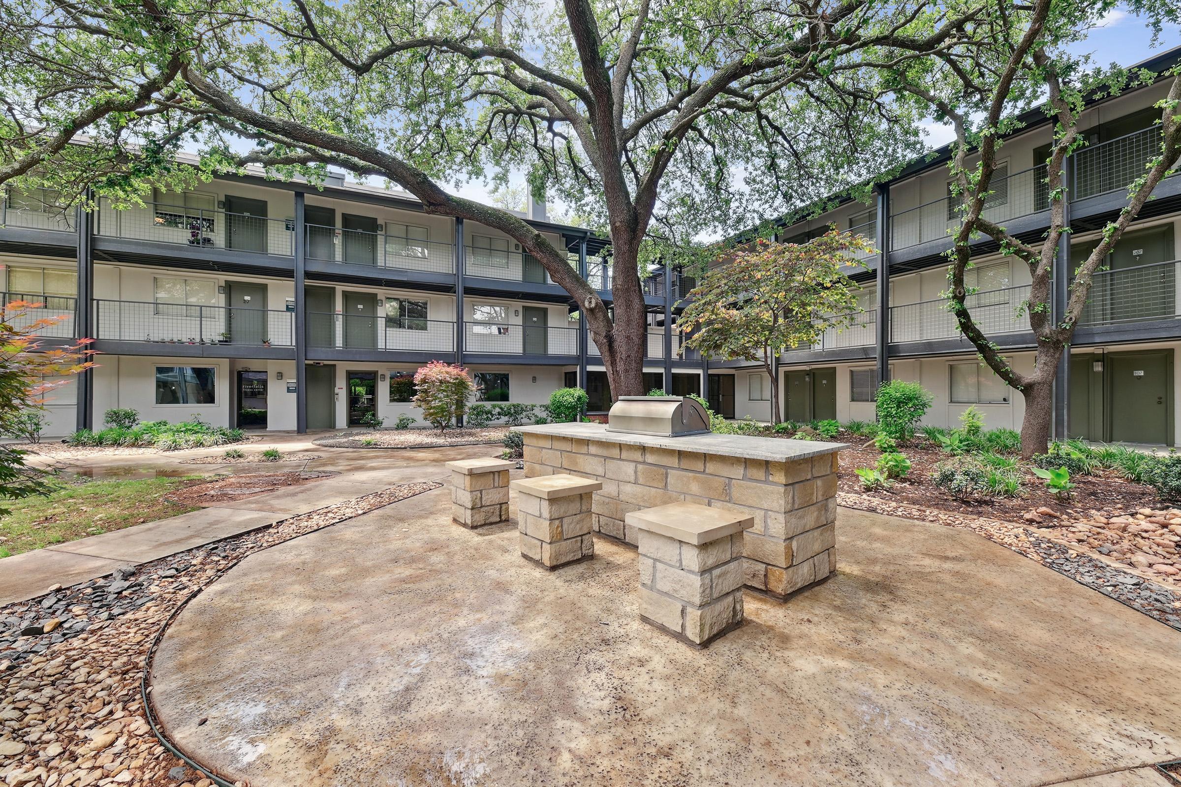 A community courtyard featuring a built-in grill and stone seating, surrounded by landscaped areas with small trees and shrubs. The backdrop includes multiple gray apartment buildings with balconies, offering a tranquil outdoor space for residents.