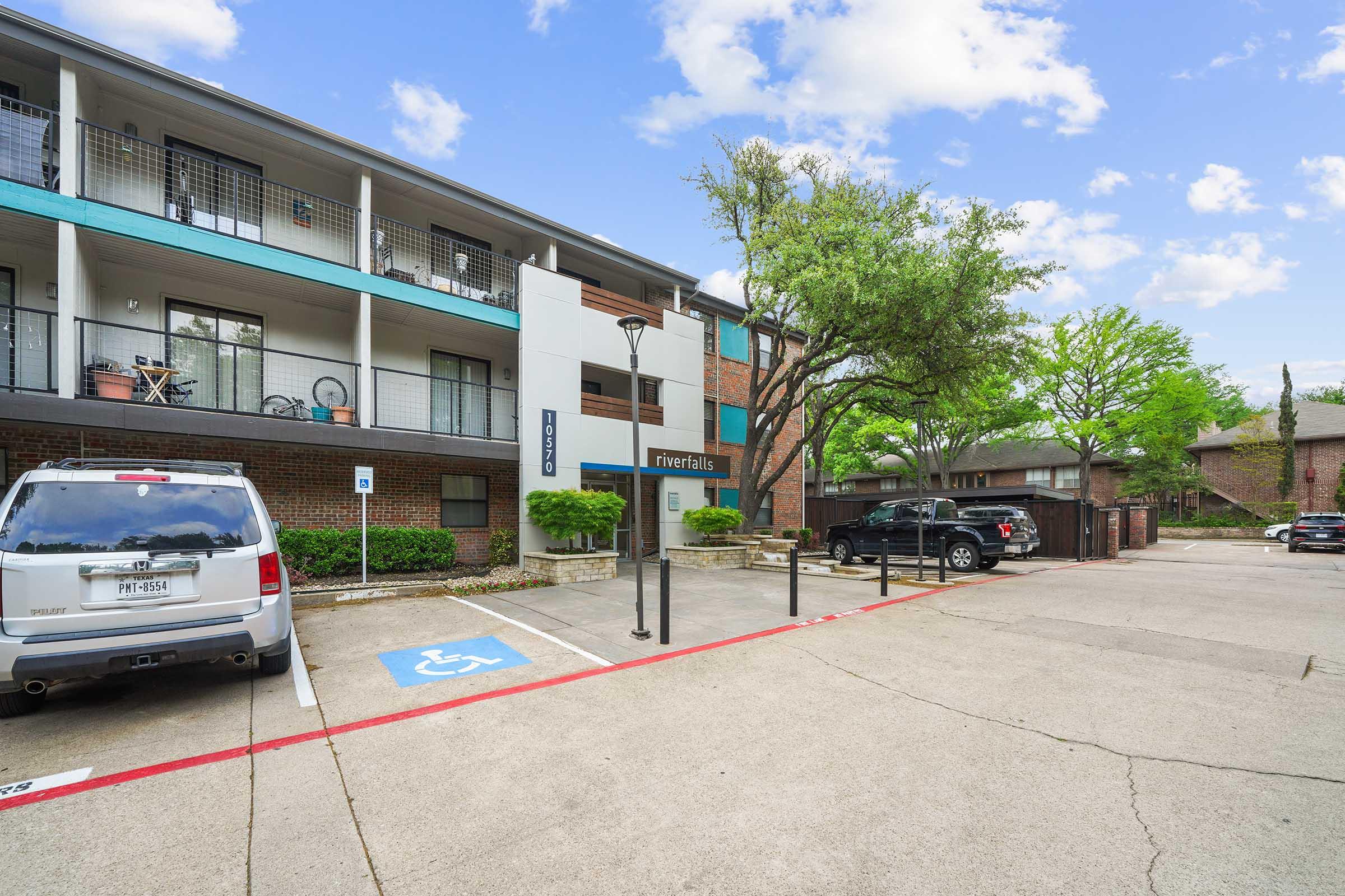 A view of an apartment complex with a parking lot. In the foreground, there is a white vehicle parked in a designated accessible space. The two-story building features balconies with plants and bicycles. Lush green trees and blue skies add to the pleasant atmosphere.
