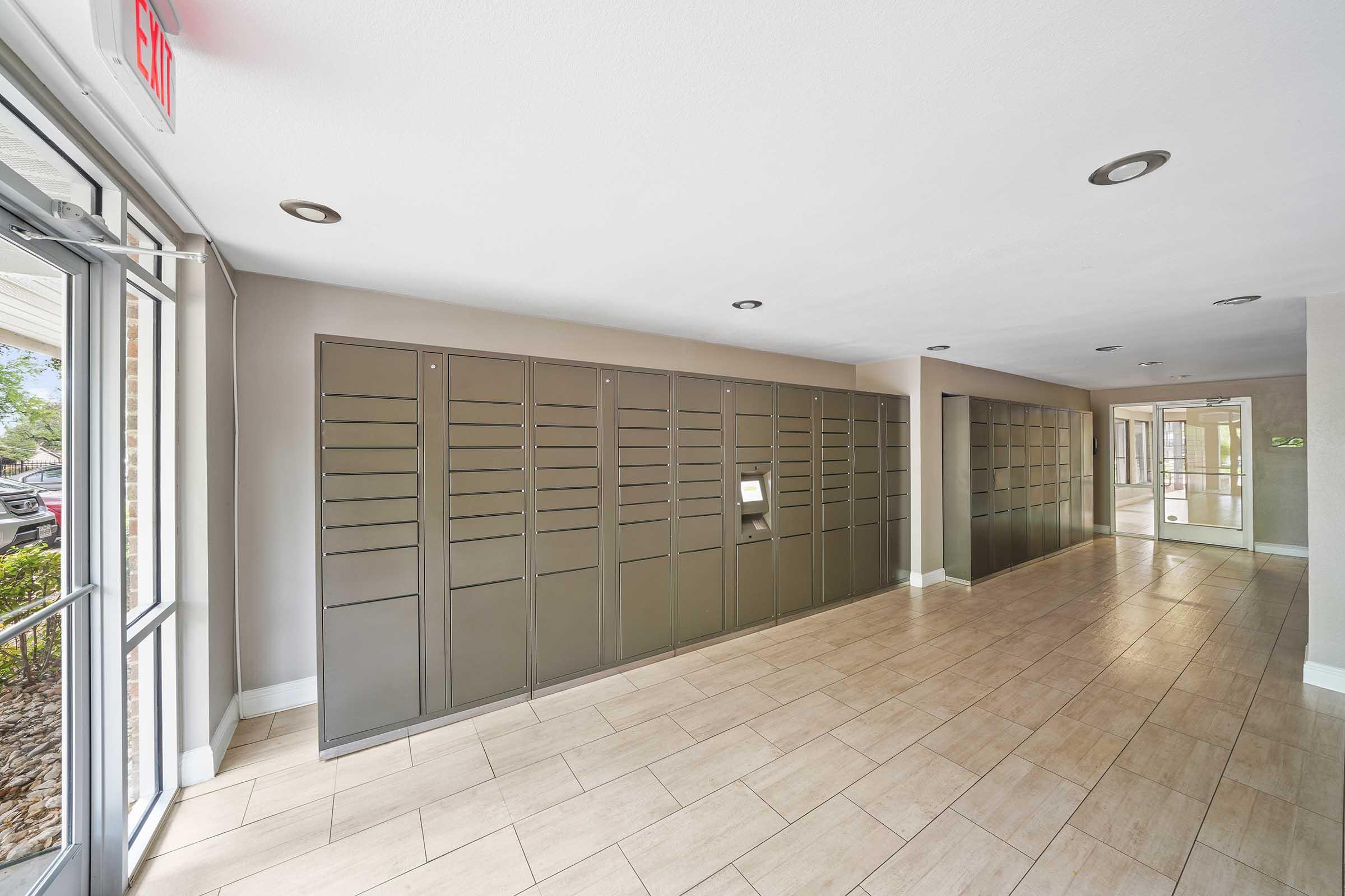 Interior view of a hallway featuring metallic mailboxes along one wall, with a clean tiled floor. The entrance is visible, showing glass doors and natural lighting. The space is well-lit and modern, designed for convenience in mail collection.