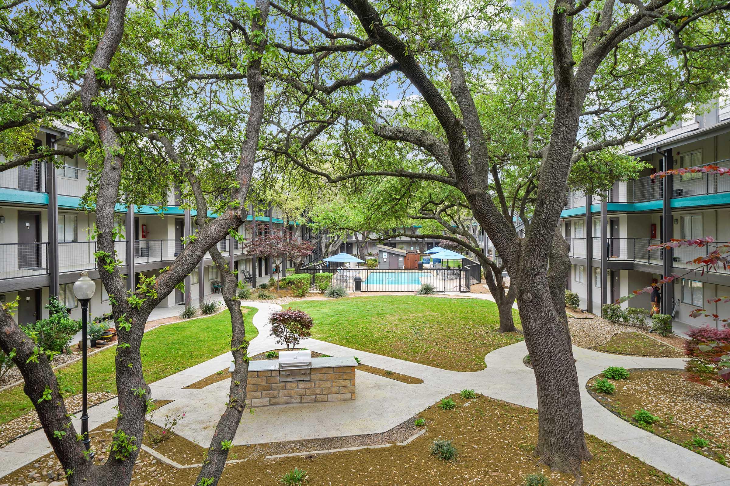 View of a landscaped courtyard in an apartment complex, featuring green grass, a central stone feature, and large trees. Surrounding the courtyard are two-story buildings with balconies. In the background, there is a swimming pool with umbrellas for shade. The scene is tranquil and inviting, with pathways leading through the greenery.