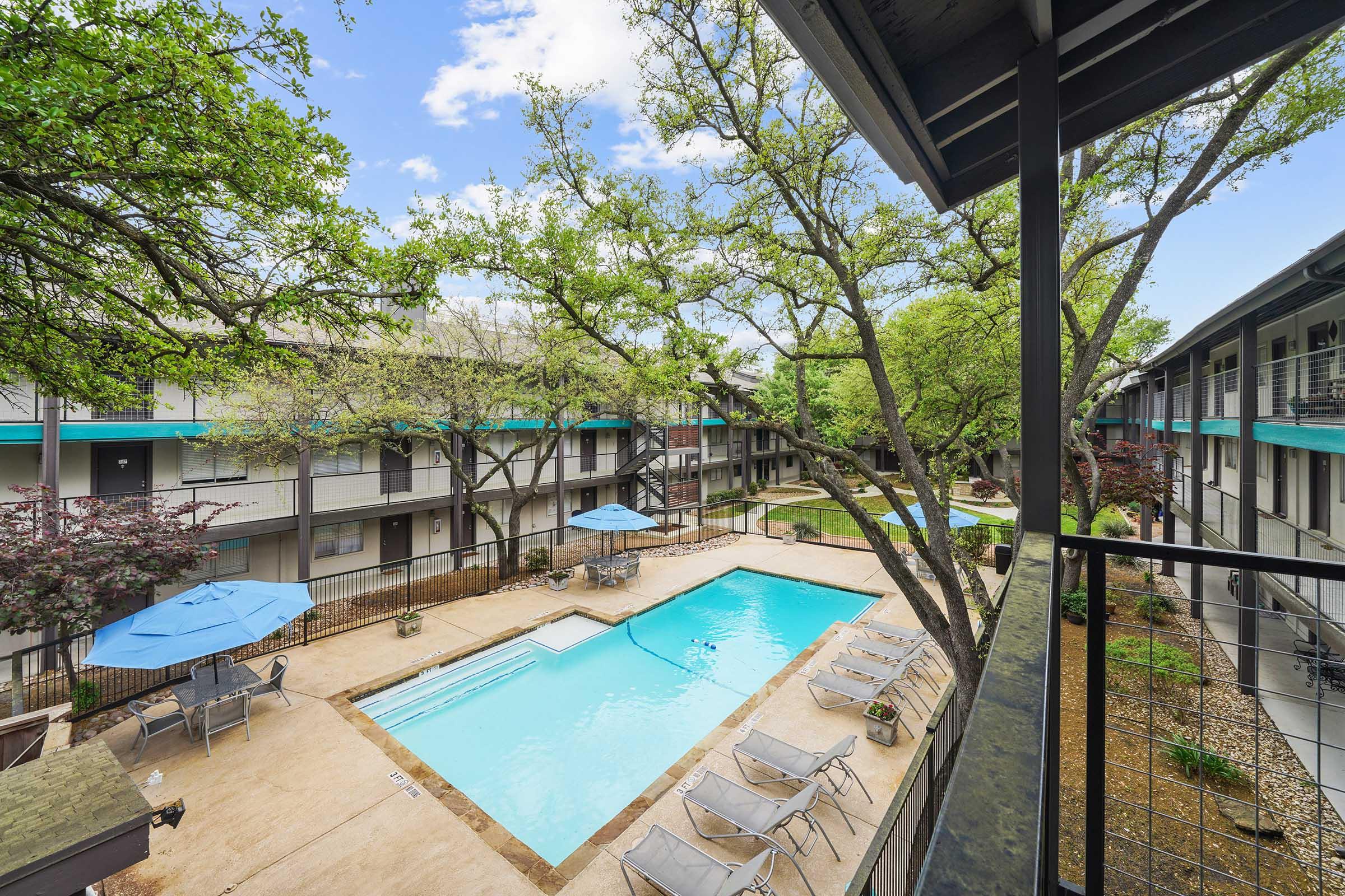 View from a balcony overlooking a pool area surrounded by trees and lounging chairs. The pool features umbrellas and is situated within a courtyard of a multi-story building. The scene is bright with blue skies and greenery, creating a relaxing atmosphere.