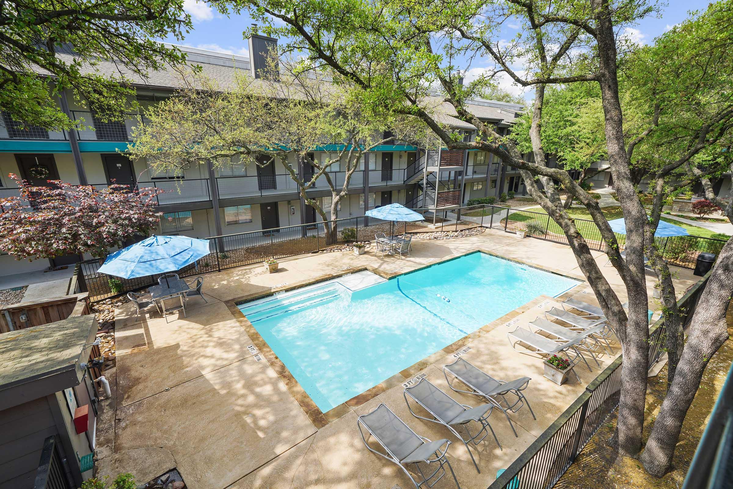 A view of an outdoor swimming pool surrounded by lounge chairs, umbrellas, and trees. The pool area is situated in front of a multi-story building with balconies and greenery, creating a relaxing atmosphere. Sunlight filters through the branches, enhancing the inviting setting.