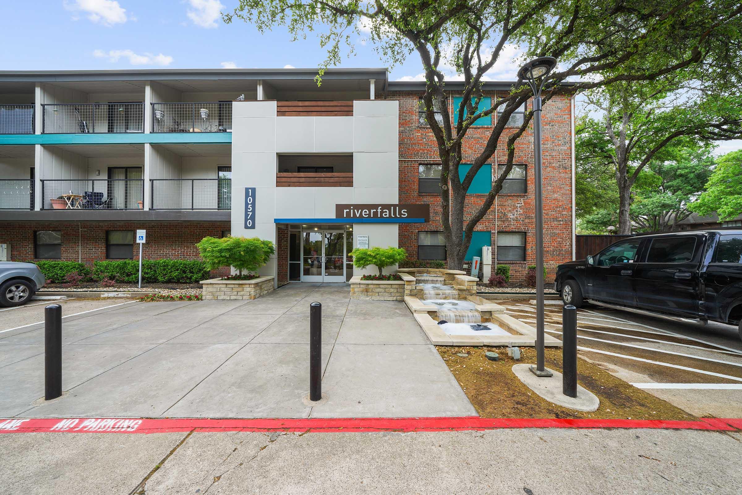 Exterior view of the Riverfalls apartment building, featuring a modern entrance with a sign labeled "riverfalls." The building has a mixture of brick and modern facade, surrounded by greenery. A parking lot is visible in front, with marked parking spaces and decorative landscaping elements.
