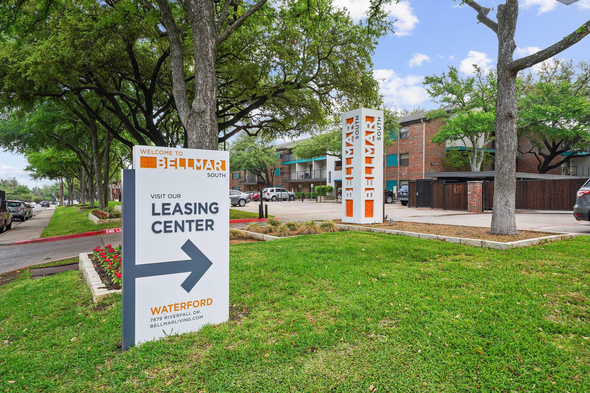 Signage directing visitors to the leasing center of a residential property, with "Welcome to Bellmar South" at the top. The sign points left, while nearby, there are well-maintained lawns and trees, and an apartment building visible in the background.