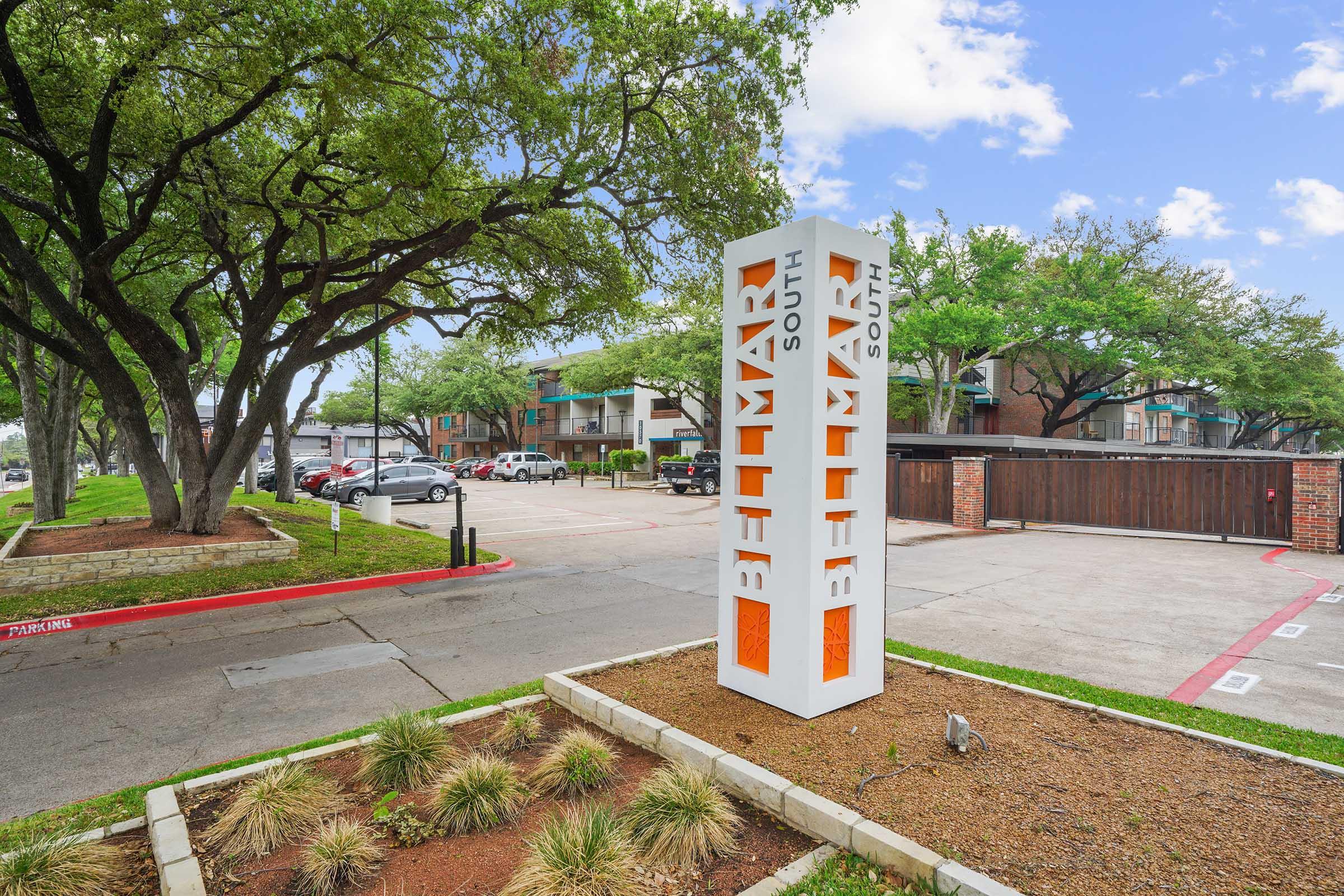 Sign for "South Lamar" with vibrant orange letters in a landscaped area. Surrounding trees and a parking lot are visible in the background, along with a multi-story building. The scene is clear and sunny, providing a welcoming atmosphere.
