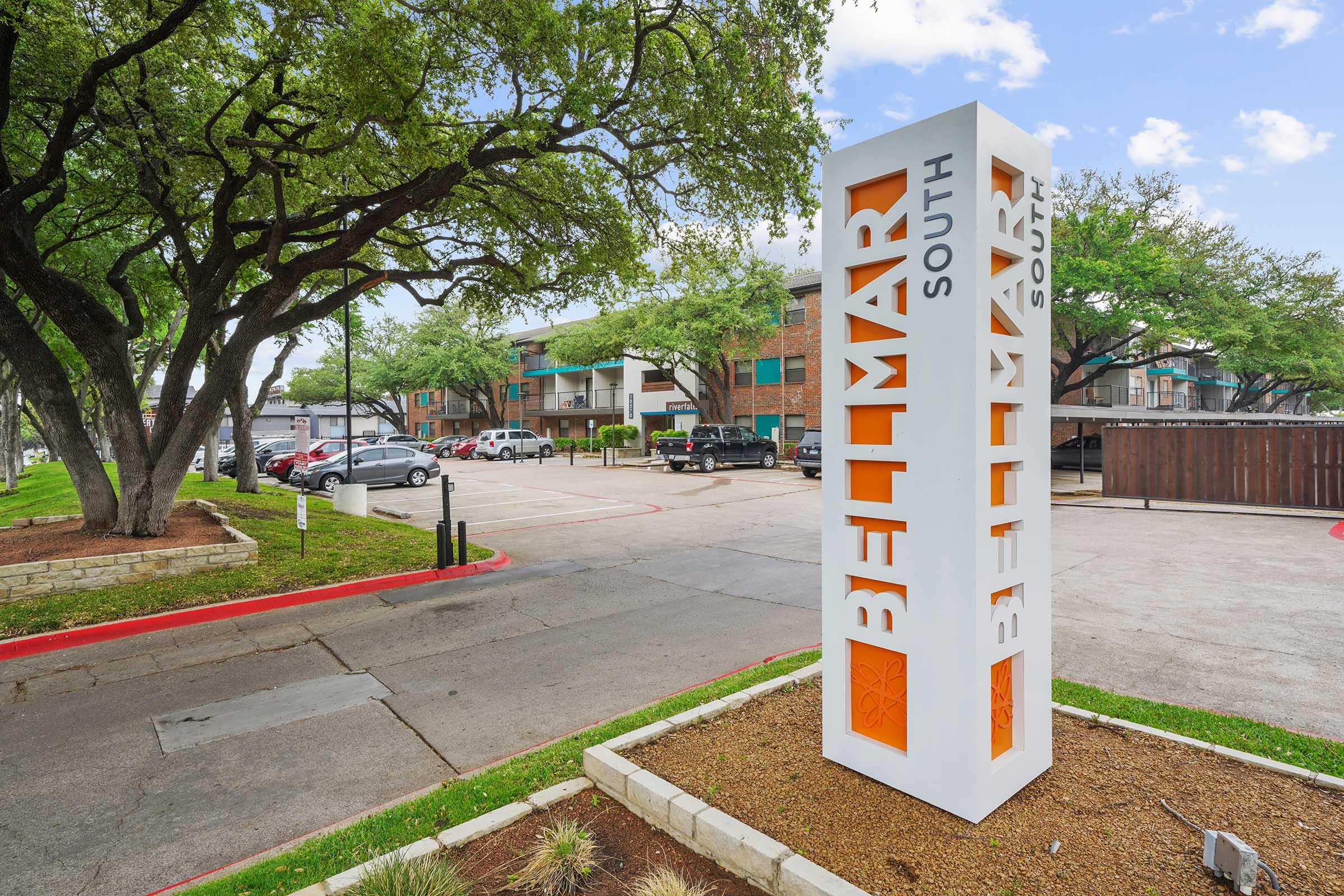 Signage for Bellmar South in a landscaped area with decorative bushes and trees. Visible parking lot with vehicles and an apartment building in the background. Clear blue sky with scattered clouds.