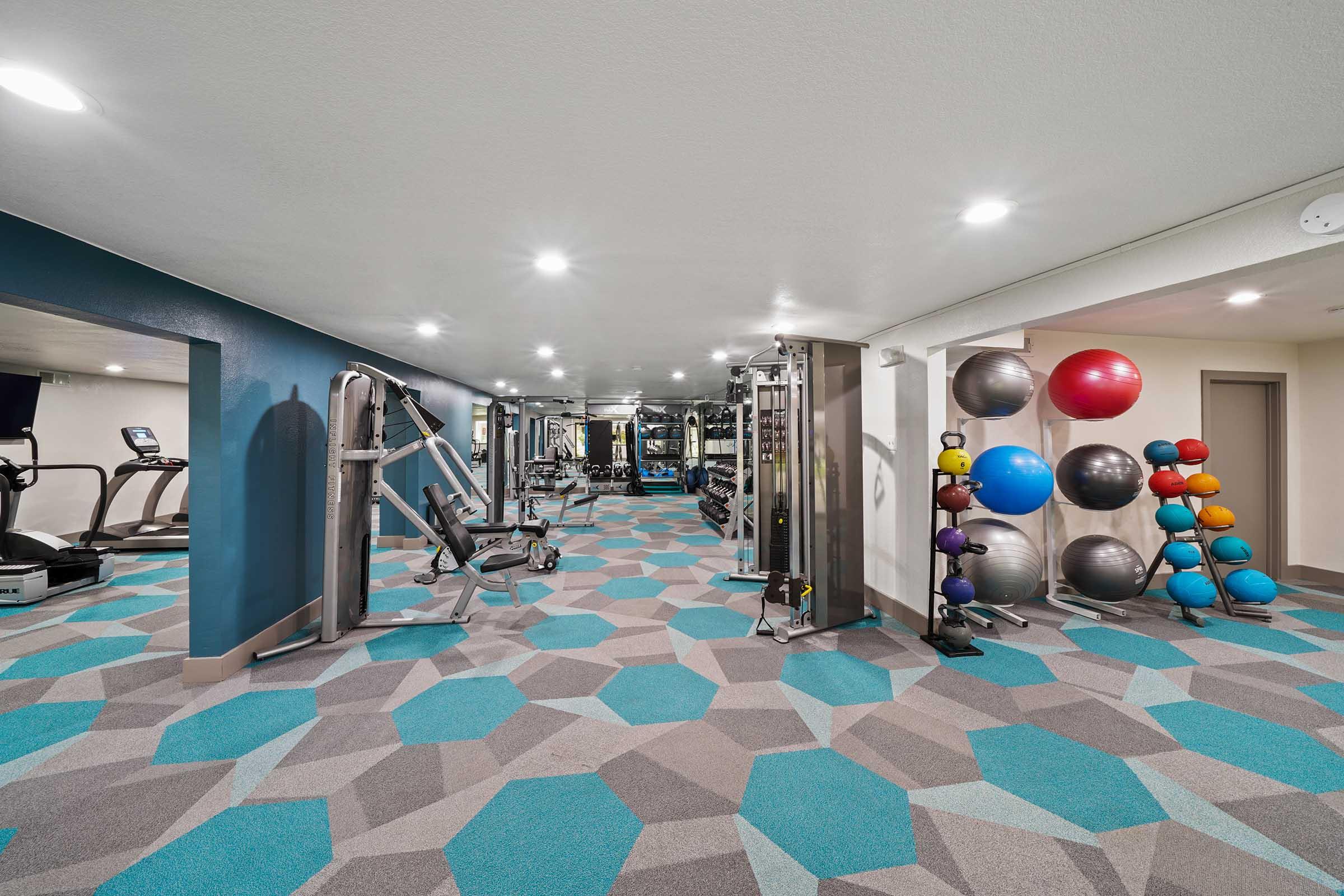A modern gym interior featuring exercise equipment such as treadmills and weight machines. The flooring has a geometric design in shades of blue and gray, and there are colorful exercise balls stacked against the wall. The space is bright with overhead lighting, creating an inviting workout environment.
