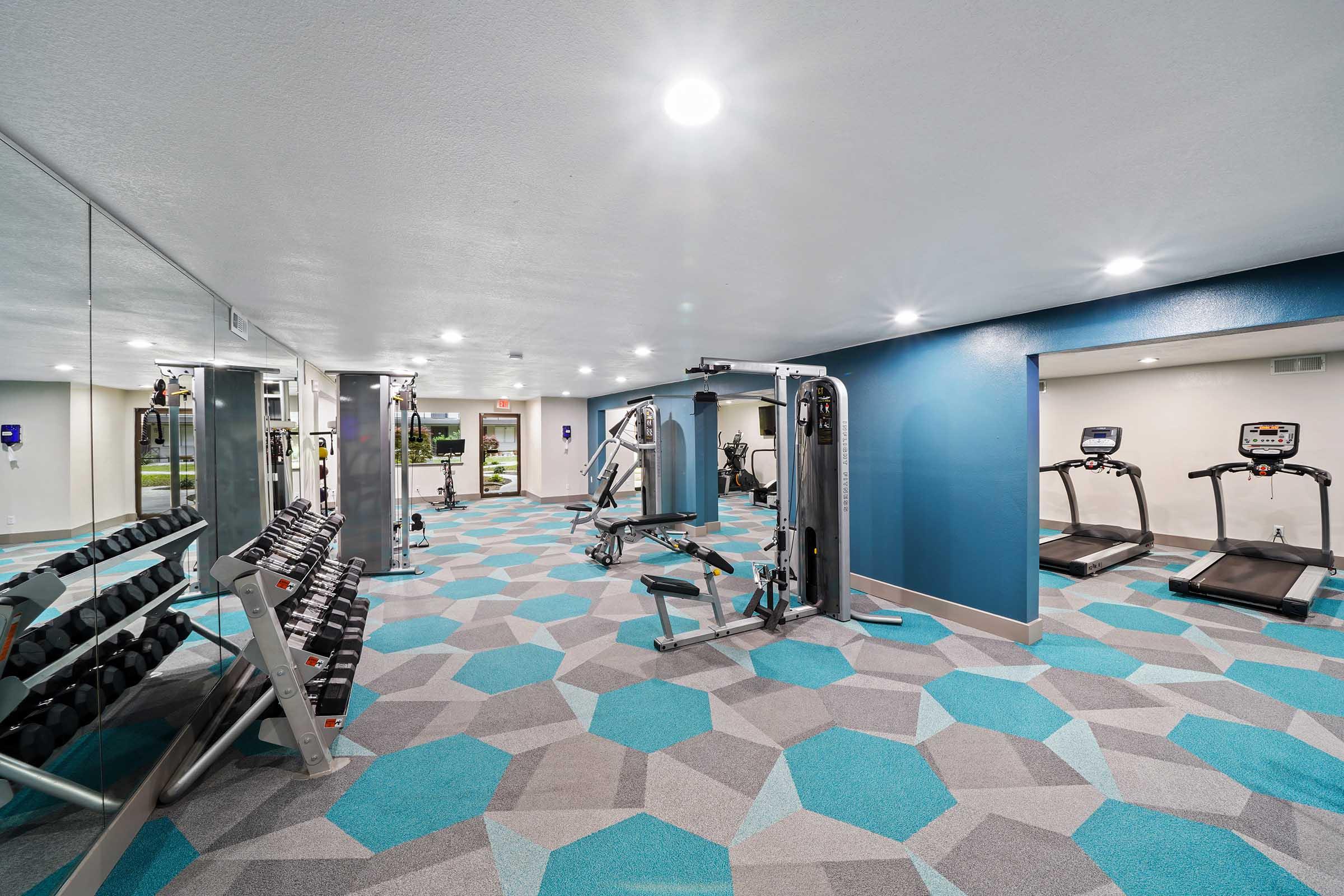 A modern gym featuring a variety of exercise equipment, including weight machines, free weights, and treadmills. The space has a bright, clean design with blue and gray hexagonal patterned flooring and mirrors, creating an inviting atmosphere for fitness activities.