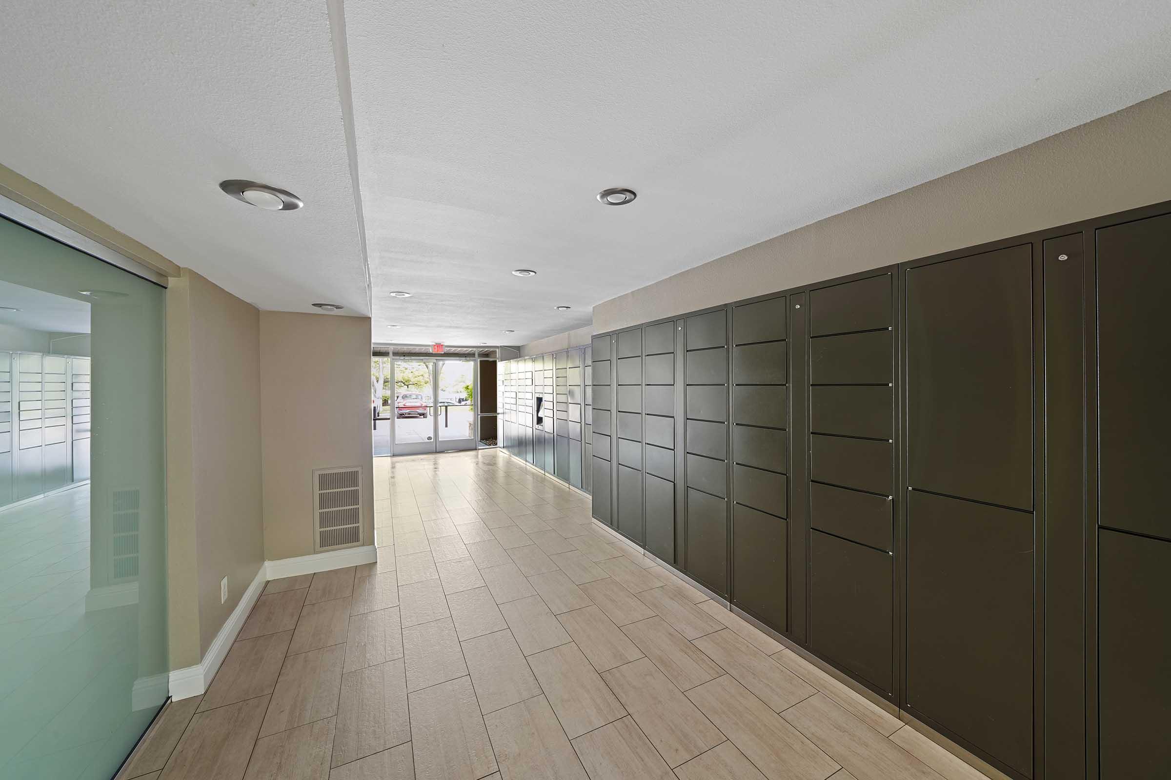 Interior hallway of a modern building featuring a row of dark green mail lockers on one side. Natural light enters through large windows at the far end, leading to an exit door. The flooring is light-colored tiles, and the walls are neutral in tone. The overall space is clean and well-lit.