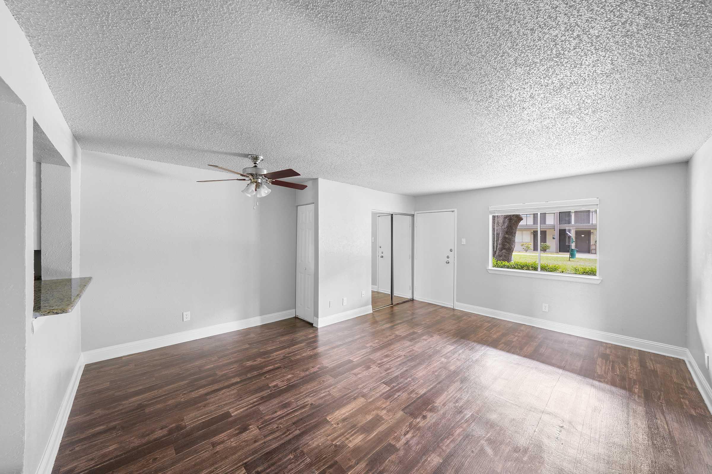 Spacious, well-lit living room featuring textured ceilings, hardwood flooring, a ceiling fan, and a large window for natural light. A door leads to another room, while a small pass-through connects to a kitchen area. Freshly painted in neutral tones, creating a cozy and inviting atmosphere.