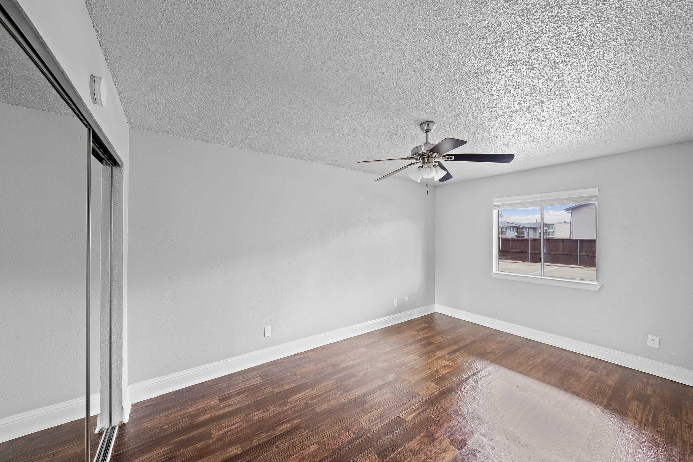 A spacious room featuring gray walls and a ceiling fan, with a window allowing natural light to stream in. The floor is covered with polished hardwood, and there's a mirrored closet on one side, adding to the open and bright feel of the space.