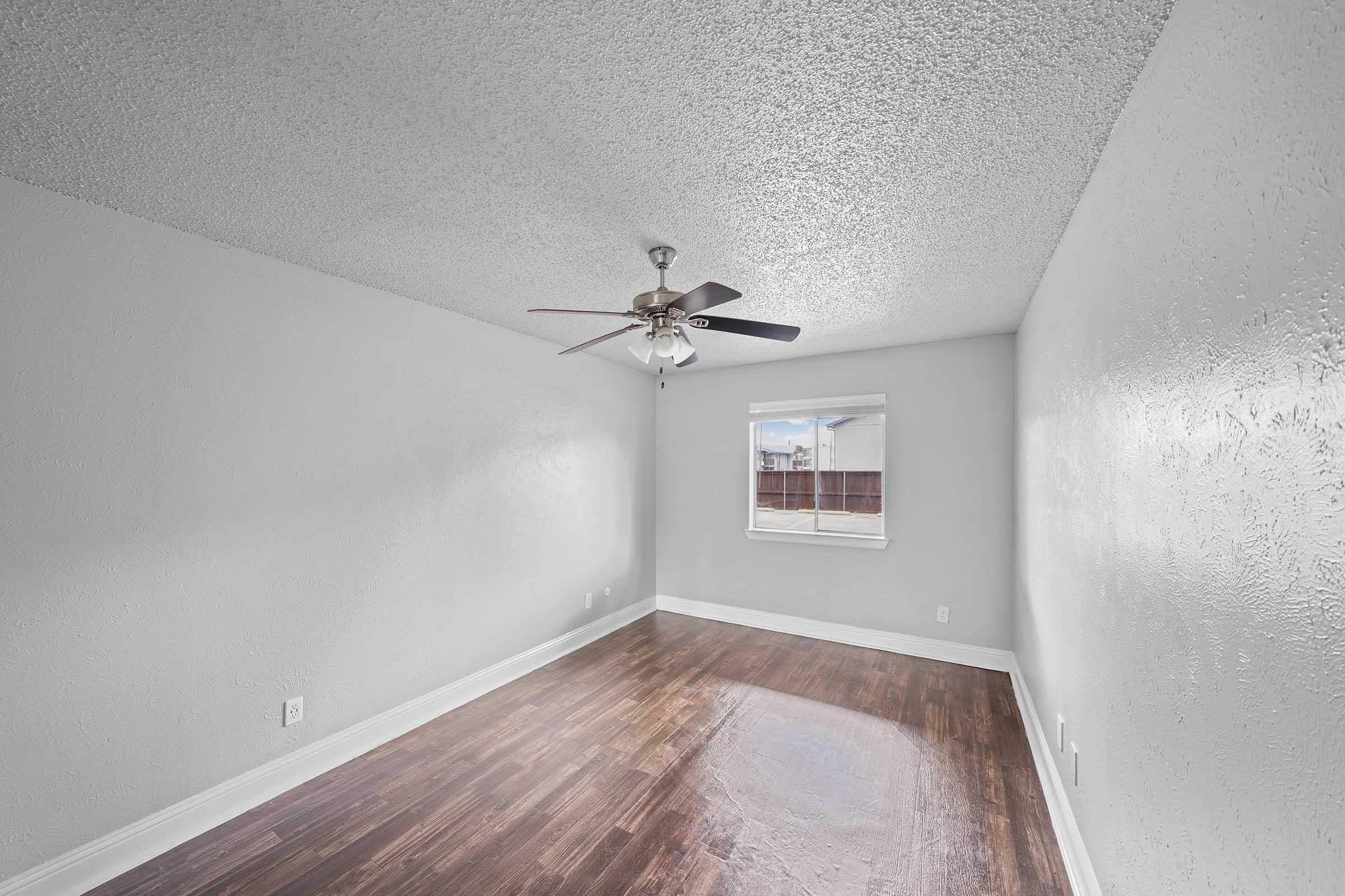 A bright, empty room with a textured white ceiling and light gray walls. The floor is dark wood laminate. A ceiling fan with dark wooden blades is visible, and a window lets in natural light, showing a glimpse of the outdoors. The overall atmosphere is clean and inviting.