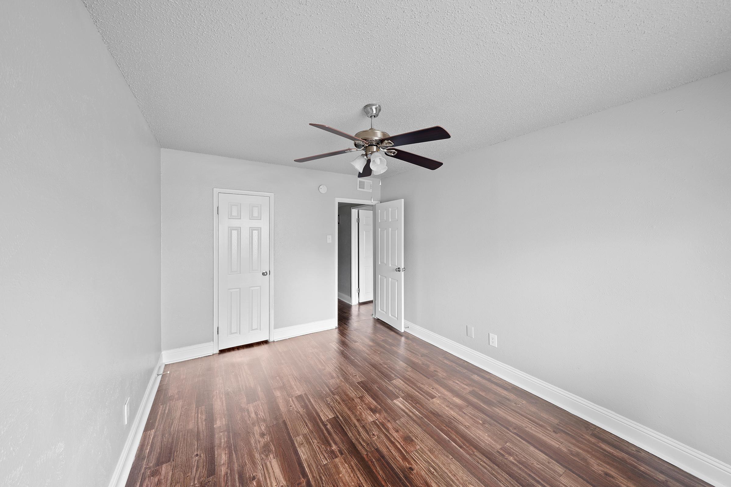 A spacious, light-colored room with a ceiling fan and hardwood floors. It features two white doors leading to other areas, and smooth gray walls. The space is empty and bright, creating a welcoming atmosphere.