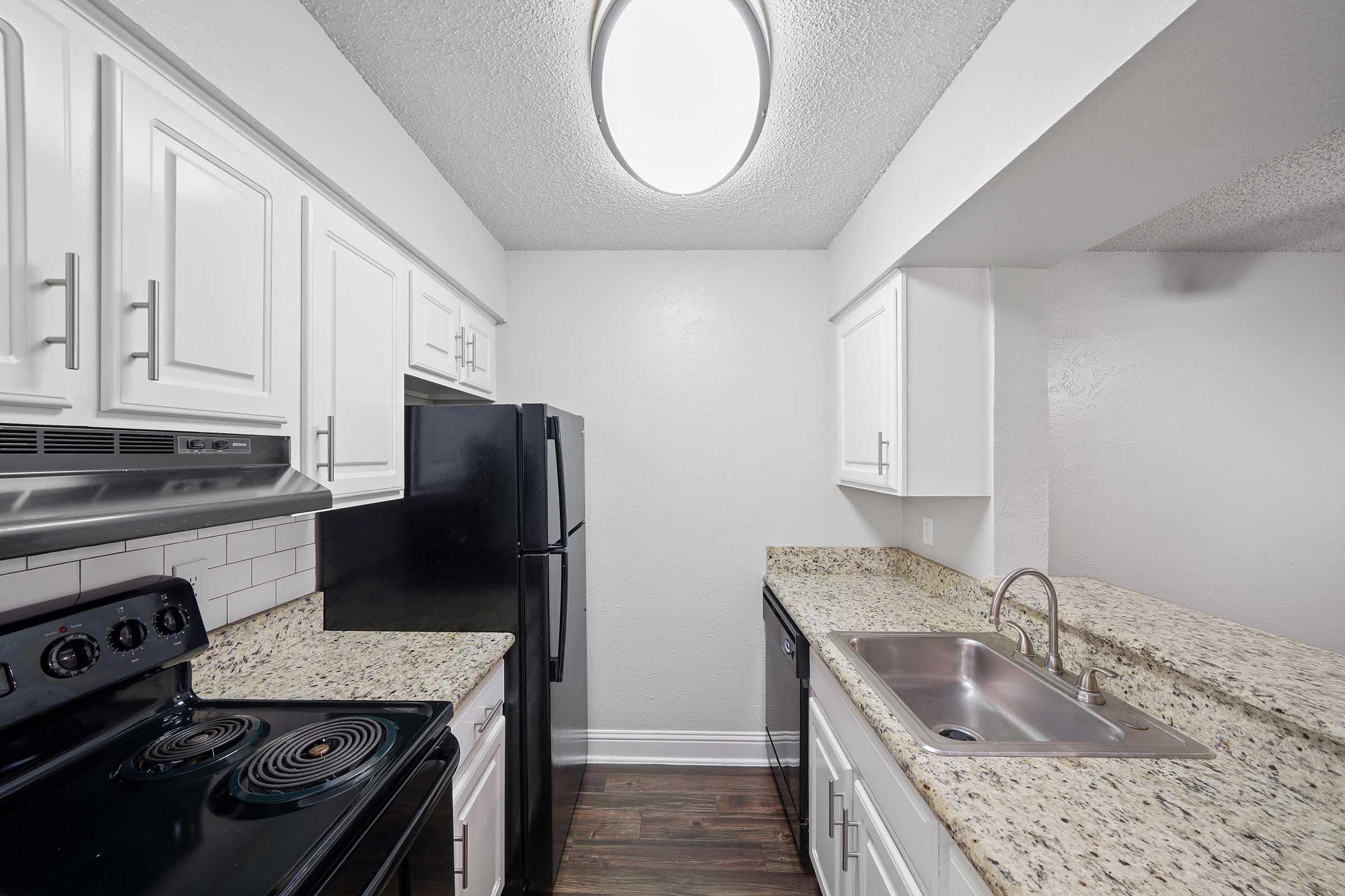 A modern kitchen featuring white cabinets, a black refrigerator, a black stove with a glass top, and a stainless steel sink. The countertop is made of speckled granite, and there's a round ceiling light providing illumination. The walls are painted a light color, creating a bright atmosphere.