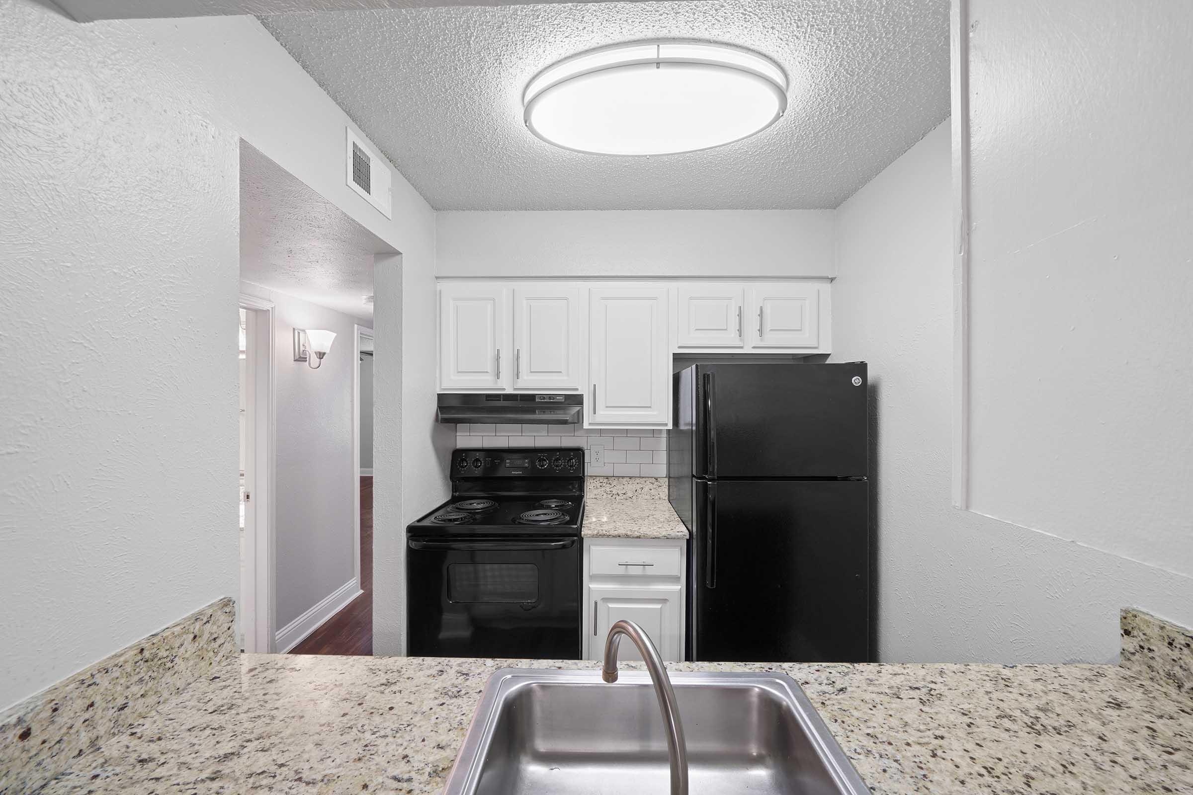 A modern kitchen featuring white cabinetry, granite countertops, a stainless steel sink, a black stove, and a refrigerator. The room is well-lit by a circular ceiling light, and there is an open view into another area of the home.