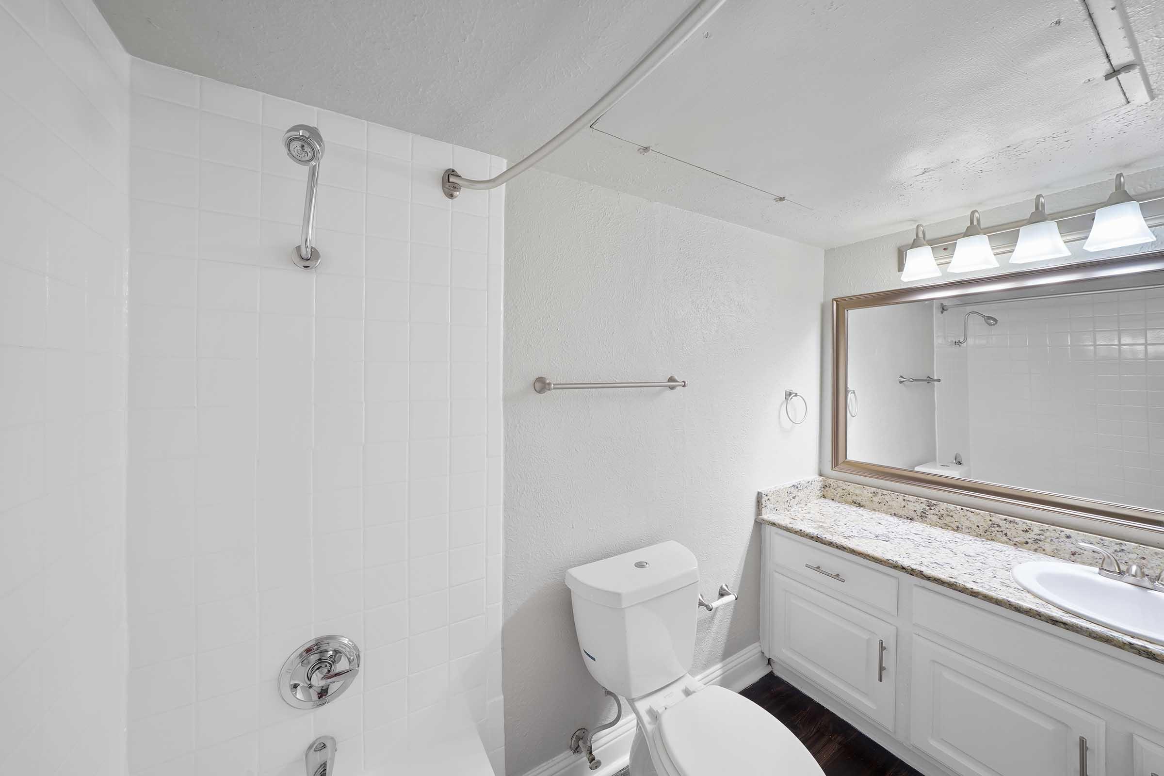A clean bathroom featuring a bathtub with a shower head, a toilet, a double sink with a granite countertop, and a large mirror above the sink. Bright lighting from three light fixtures is installed above the mirror, while the walls are primarily white with a neutral tile design.