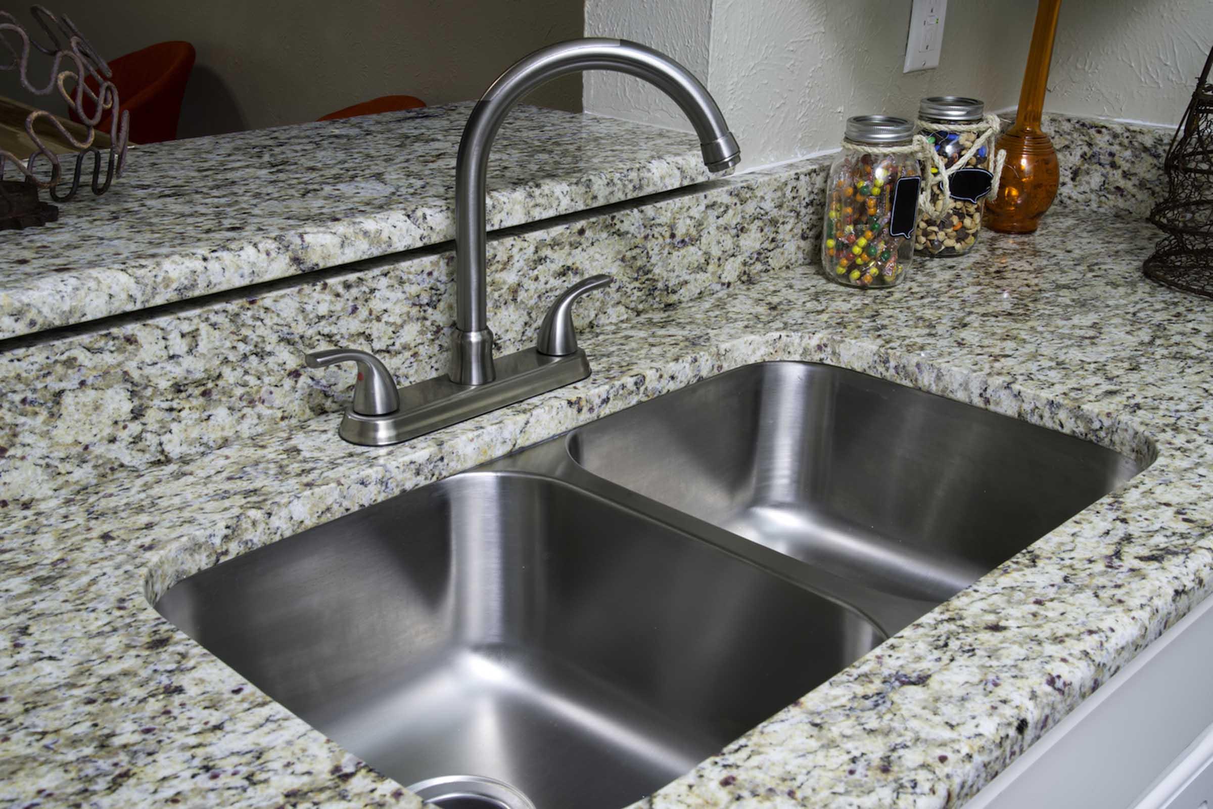 A stainless steel double sink sits on a speckled granite countertop. A modern faucet is positioned above the sink. In the background, there are jars filled with colorful candies and nuts, along with a decorative orange bottle, adding a touch of decor to the kitchen space.