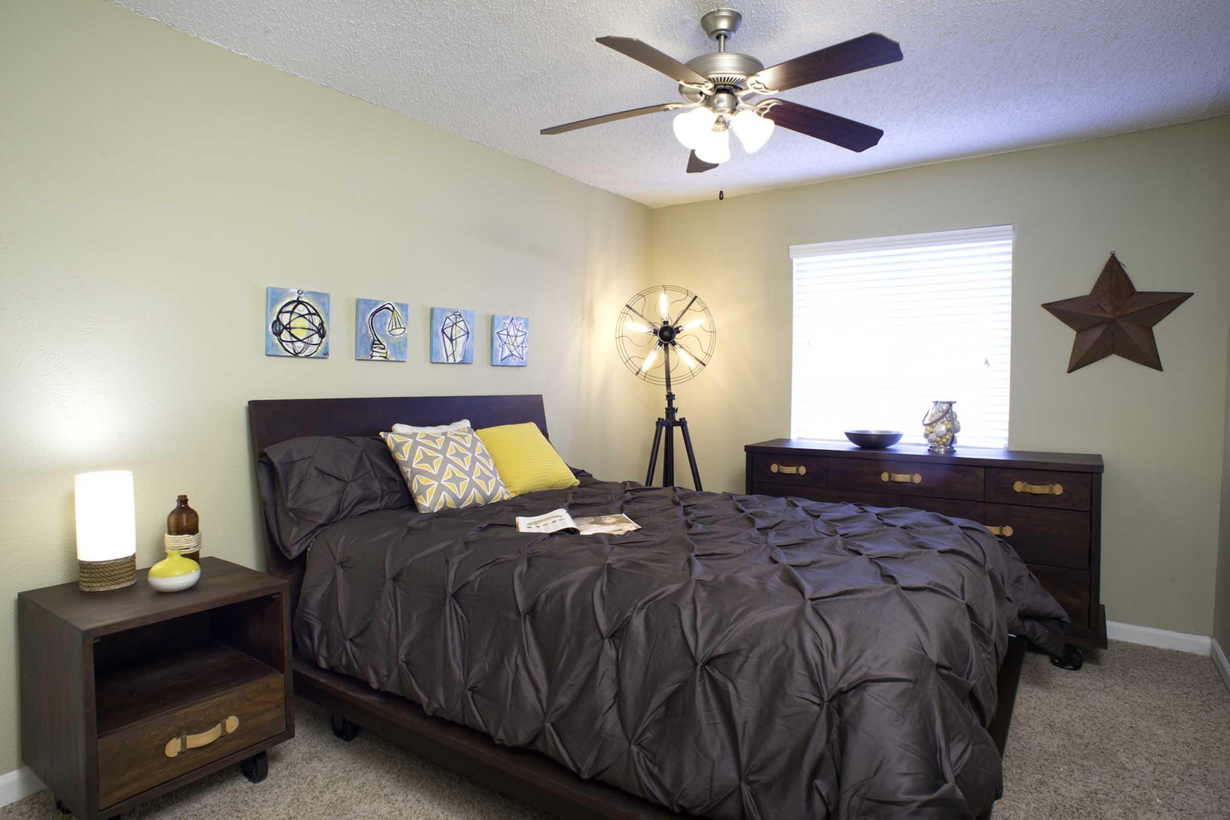 A cozy bedroom featuring a queen-sized bed with a dark comforter and yellow accent pillows. There are a bedside table with a lamp and a decorative bottle, along with a matching dresser. The wall is adorned with framed artwork, and a window allows natural light to brighten the room. A star-shaped decor piece enhances the aesthetic.