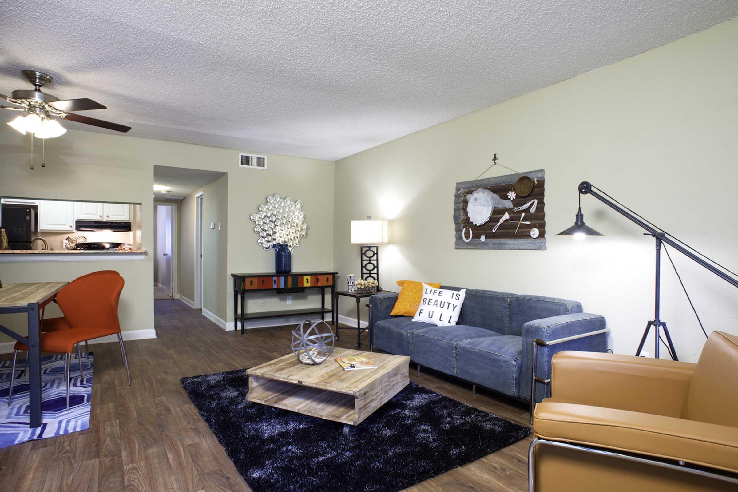 A modern living room featuring a blue sofa, an orange armchair, and a wooden coffee table on a black rug. There's a ceiling fan, a decorative light fixture, and a wall art piece. In the background, a dining area is visible with a wooden table and colorful chairs. Neutral-colored walls enhance the inviting atmosphere.