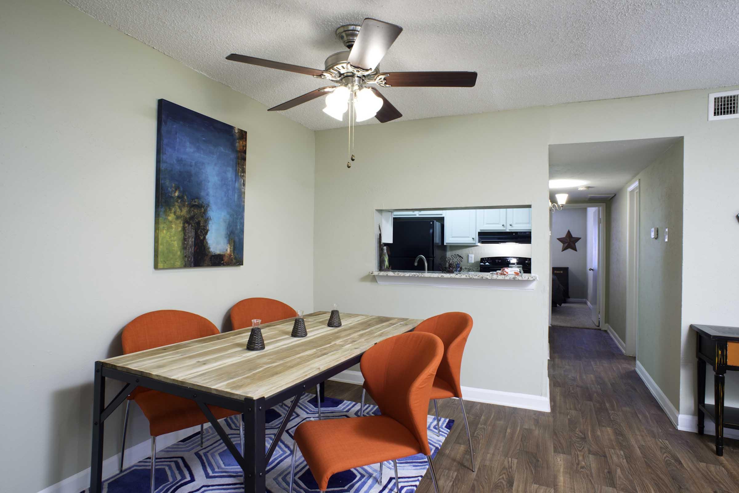 A modern dining area featuring a wooden table with four bright red chairs. The walls are painted in a light color, and there's a colorful abstract painting on one wall. A ceiling fan is visible, and the space opens into a kitchen area in the background. A hallway leads to another room, adding depth to the layout.