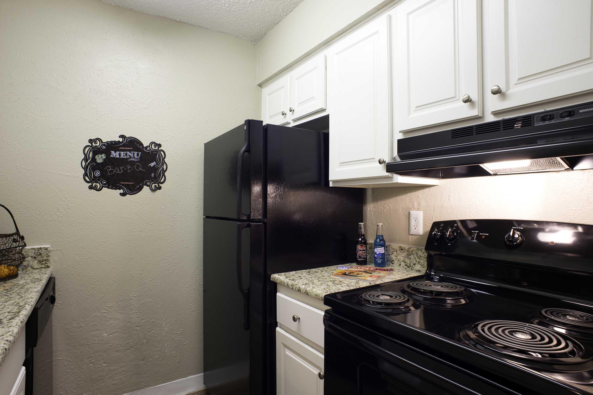 A modern kitchen featuring white cabinets, a black refrigerator, and a black stove with a granite countertop. A decorative sign labeled "MENU" is mounted on the wall, and two bottles are displayed on the countertop alongside a pizza. The overall aesthetic is bright and inviting.