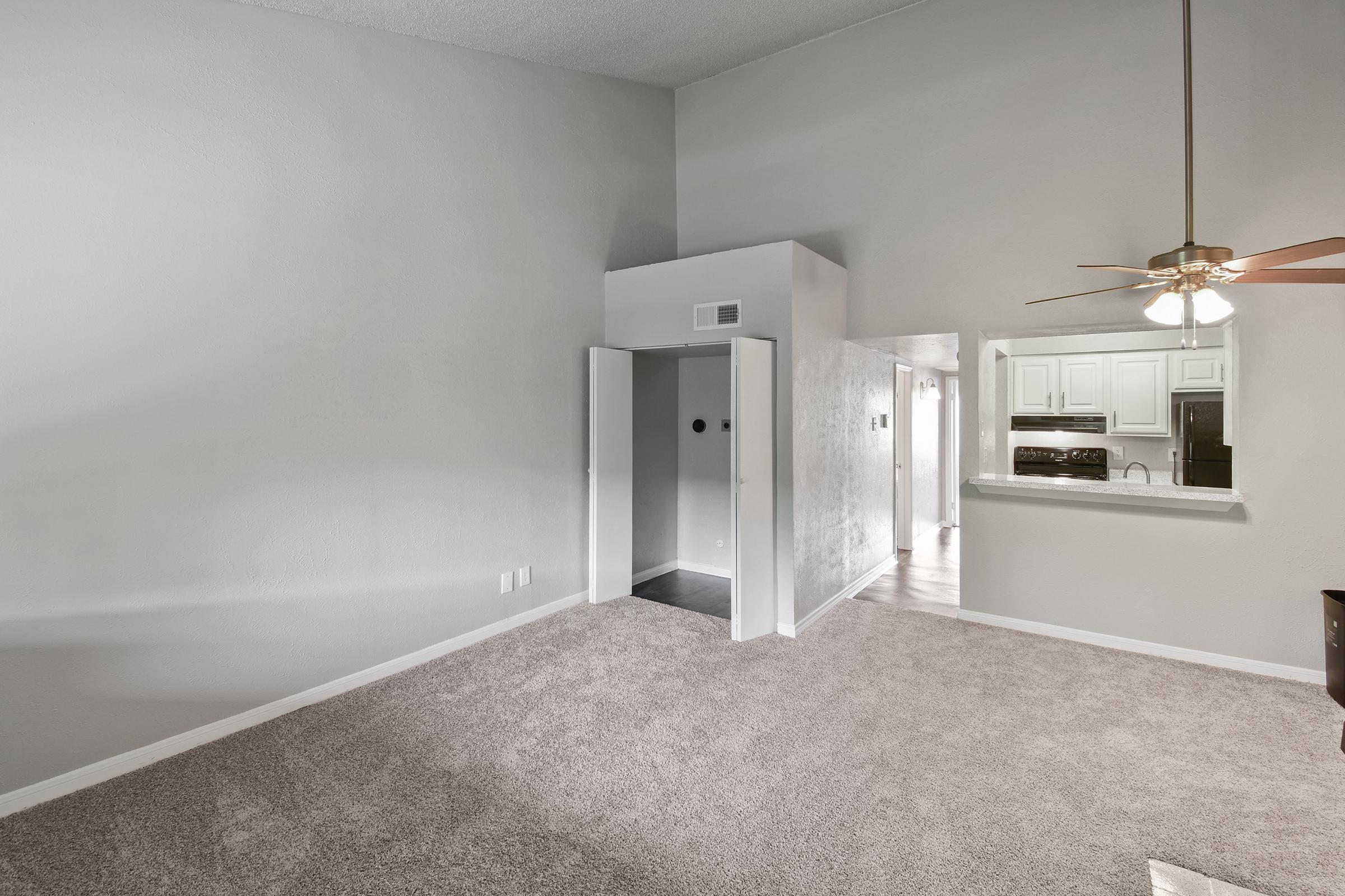 Interior view of an apartment featuring beige carpet, a high ceiling, and light gray walls. A doorway is visible on the left, leading to a kitchen area with white cabinets in the background. A ceiling fan is installed in the corner, and there is a sense of spaciousness in the room.