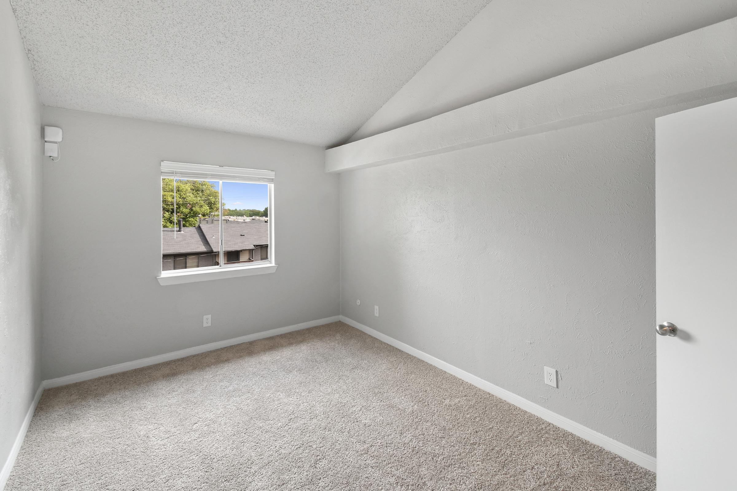 A well-lit, empty room with beige carpet and light gray walls. It features a sloped ceiling, a single window with a view of nearby buildings, and a white door. The space feels clean and minimalistic, perfect for a living area or bedroom.