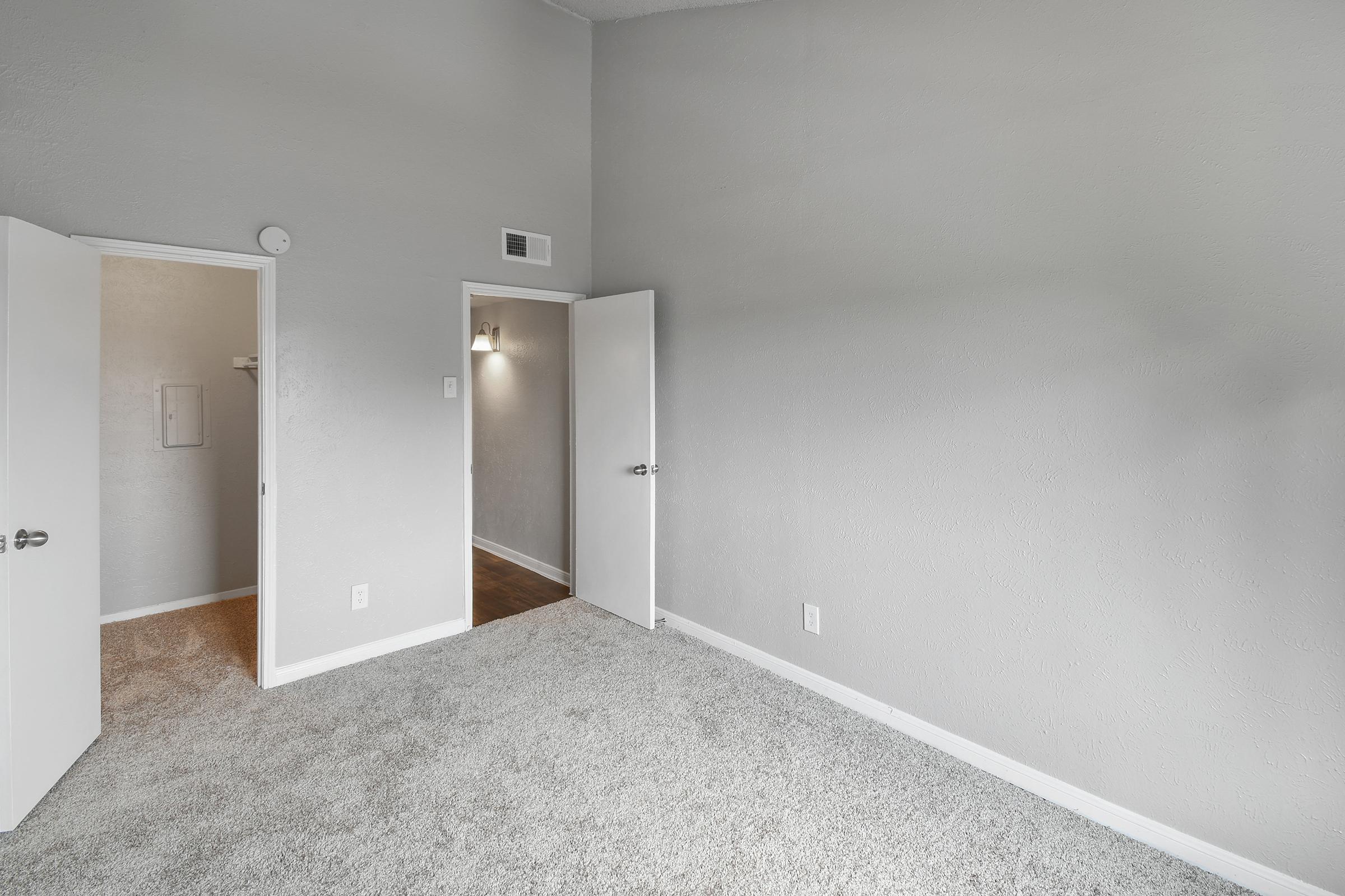 A spacious room with light gray walls and carpeted flooring. Two doors lead to other areas, one possibly a closet and the other to a hallway. Soft lighting enhances the modern, airy feel of the space, which is empty and ready for personal customization.