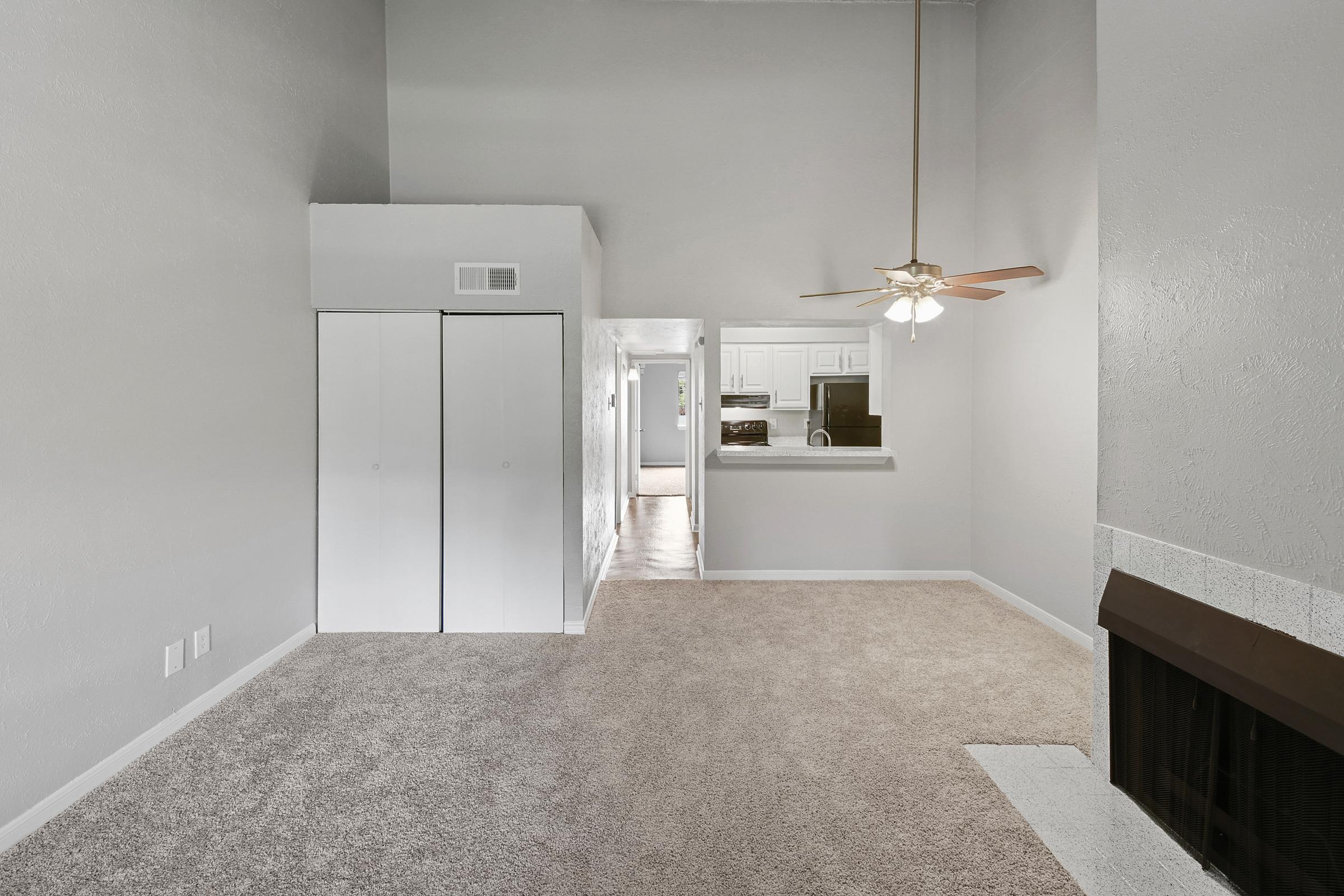 Spacious living area with neutral gray walls and plush carpet. Features a ceiling fan and a decorative fireplace. Two white closet doors on the left and an open view toward the kitchen in the background. Bright, inviting atmosphere with natural light.