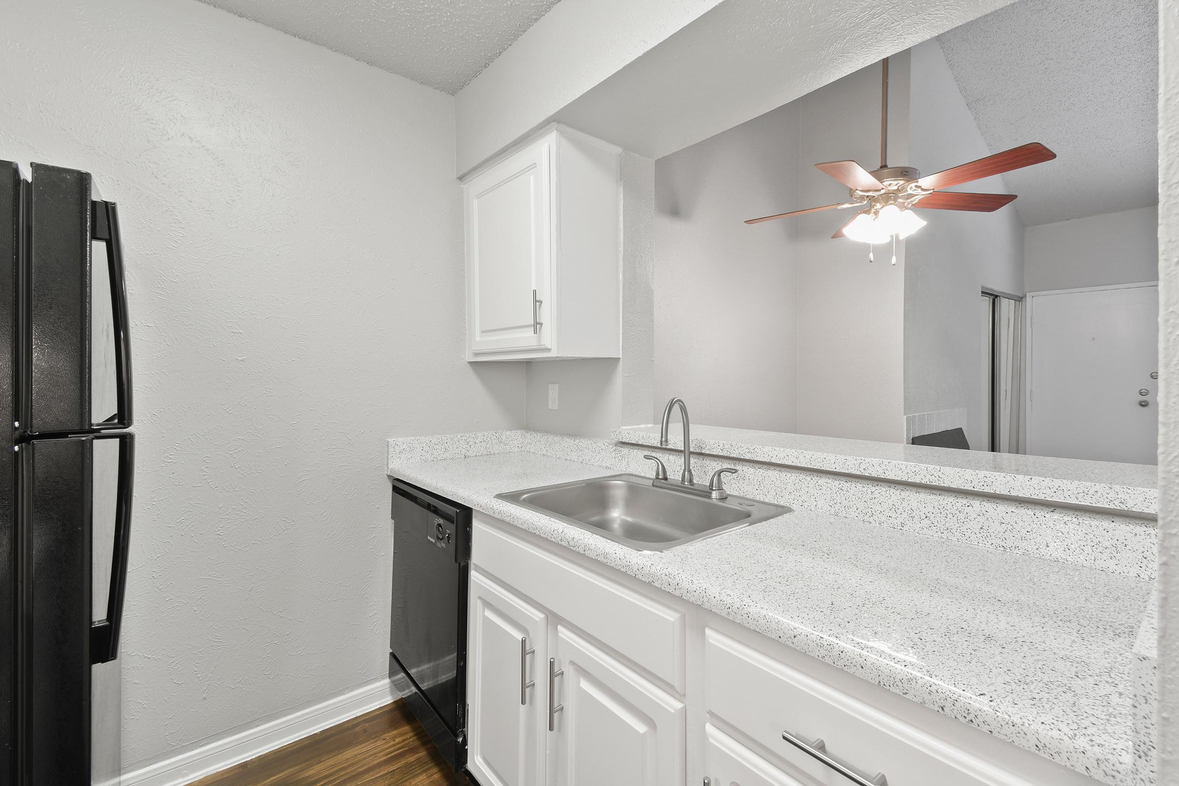 A modern kitchen featuring white cabinetry, a black refrigerator, a black dishwasher, and a double sink. The countertop is light-colored with a speckled design. A ceiling fan with red blades is visible above, and the walls are painted in a light shade, contributing to a bright and airy atmosphere.