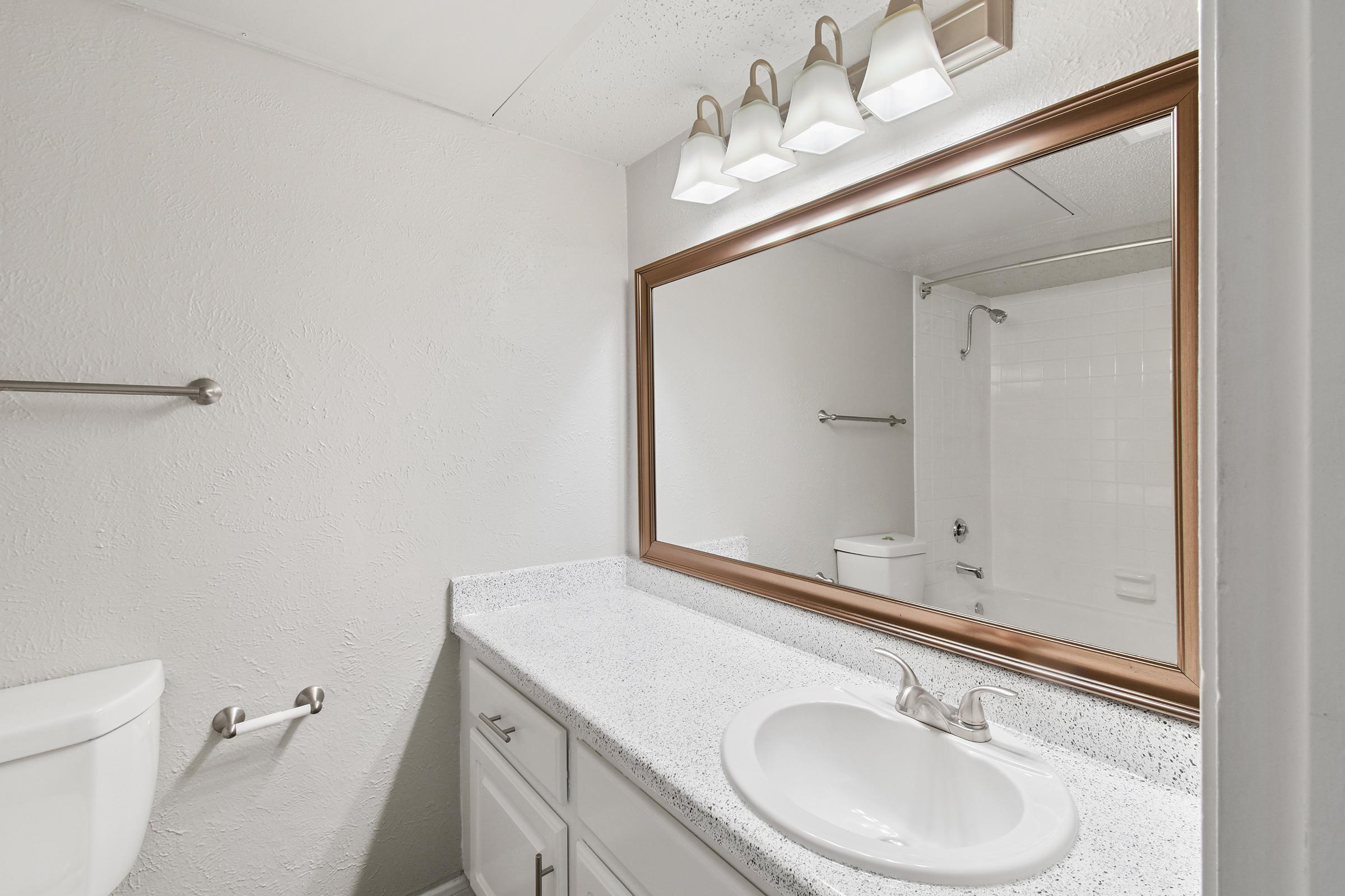 A clean, modern bathroom featuring a white sink with a chrome faucet, a large mirror with a bronze frame, three light fixtures above, and a white tiled shower in the background. The walls are painted light gray, and there is a towel bar on the left side. A toilet is visible to the right.