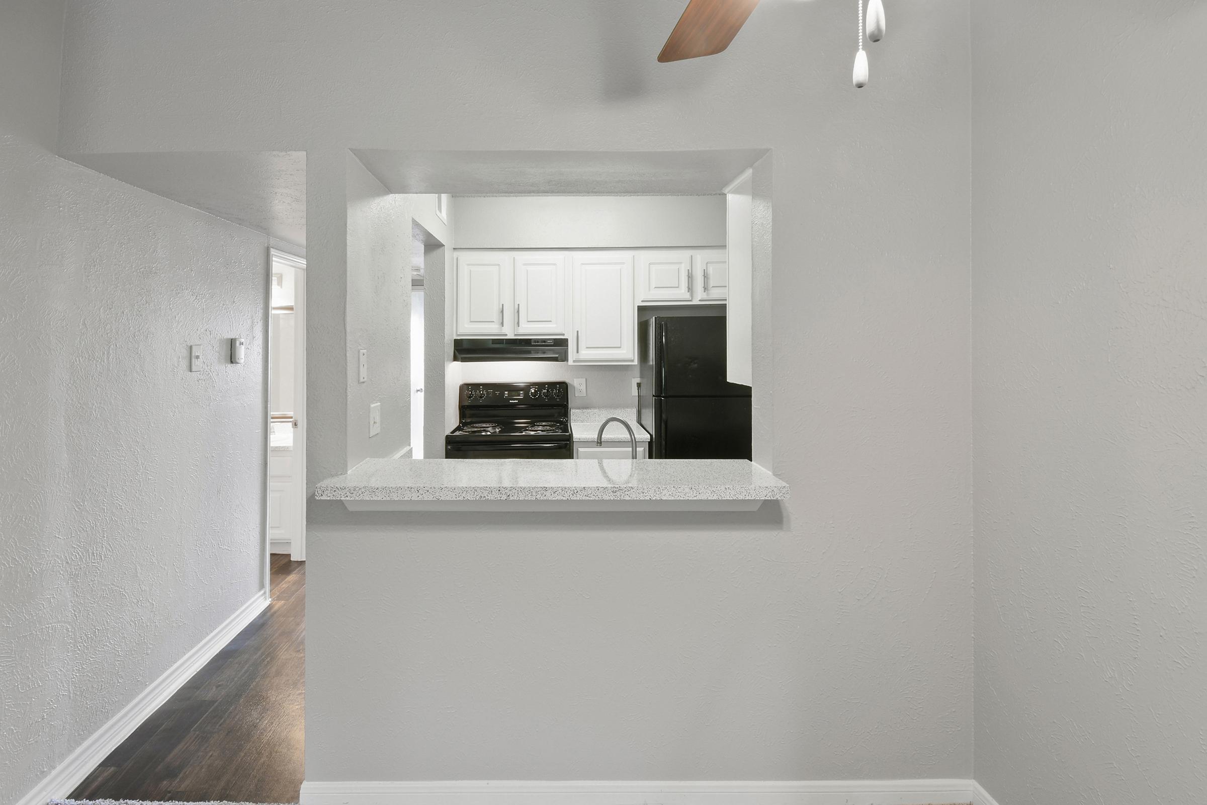 A modern, open-concept kitchen seen from a living area. The kitchen features white cabinetry, a black stove, and a refrigerator. A large window-like opening allows visibility between the rooms, with a light-colored countertop and a ceiling fan above. The walls are painted in soft gray tones.