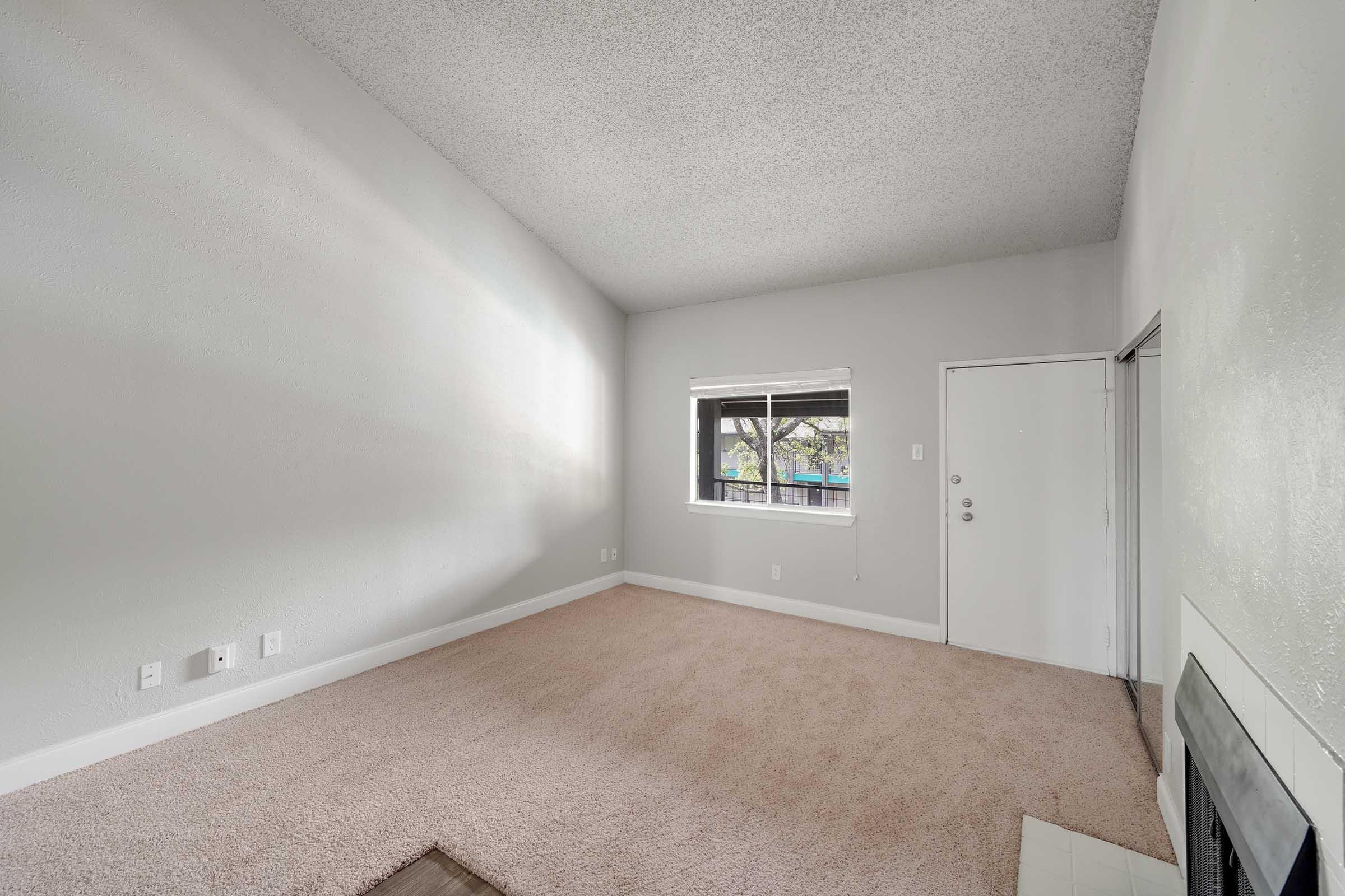 A spacious, empty room featuring light-colored walls and plush beige carpeting. There is a window on one side allowing natural light in, and a door leading to the exterior. The room has a high ceiling and a simple fireplace along one wall, creating a cozy ambiance.