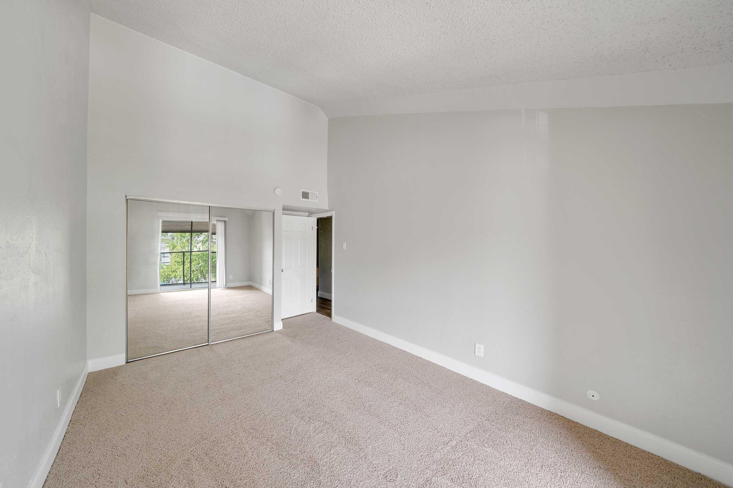Spacious room with light gray walls and carpet flooring. Features a large window with a view and a mirrored closet door. The ceiling is high, creating an airy atmosphere. The layout is open, providing a versatile space suitable for various furnishings.