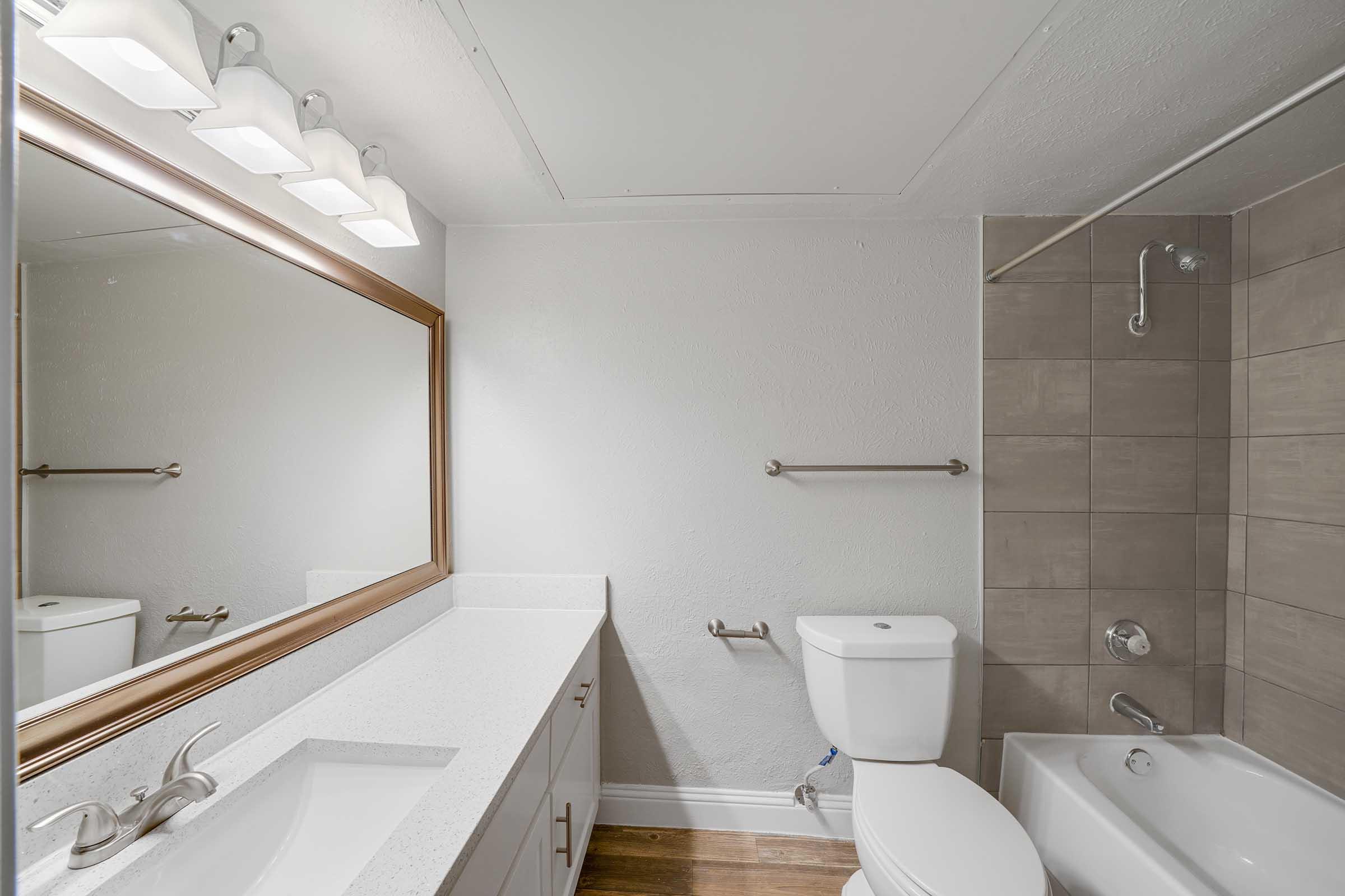 A modern bathroom featuring a white countertop with a large mirror above, a sleek faucet, a toilet, a bathtub with a showerhead, and light gray tiled walls. Soft lighting from three fixtures above the mirror complements the neutral color scheme of the space.