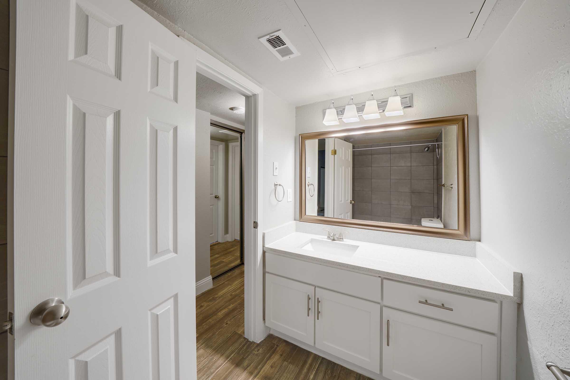 A modern bathroom featuring a white vanity with a sink, a large mirror above it, and pendant lighting. The walls are painted light colors, and there's a gray tiled shower area visible in the background. The floor is a wood-like laminate, and there's a door leading to another room.