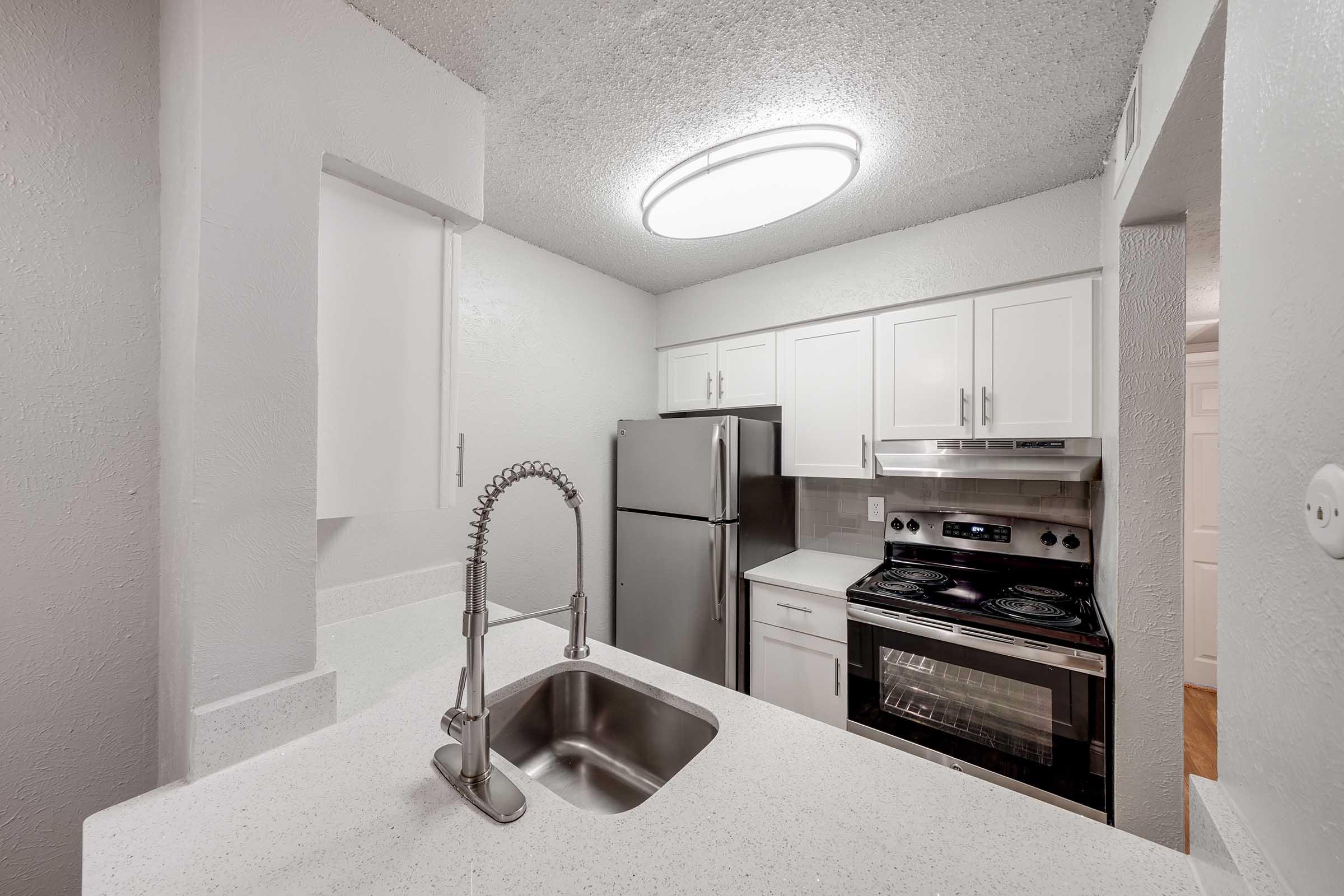 A modern kitchen interior featuring white cabinets, a stainless steel refrigerator, and an electric stove. The countertops are light-colored, and the space is well-lit by a round ceiling light. A sleek sink with a modern faucet is visible, creating a clean and contemporary atmosphere.