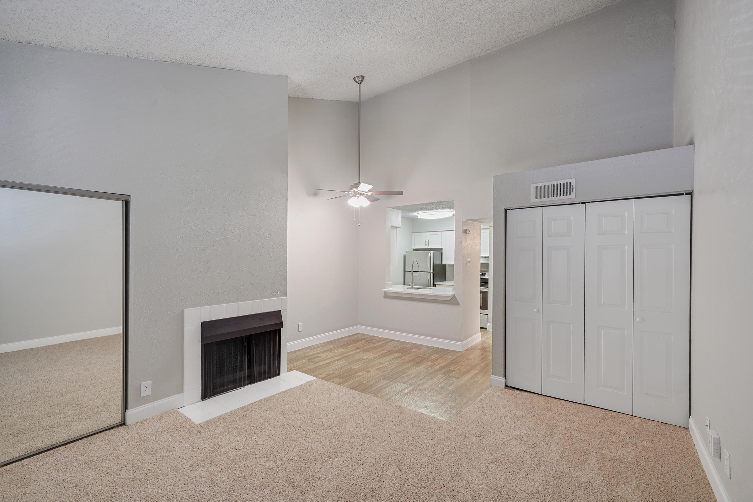 A spacious living room with light gray walls, a ceiling fan, and a white fireplace. There is a large mirror on the left and a doorway leading to a kitchen area in the background. The room features beige carpet flooring and a closet with white sliding doors.