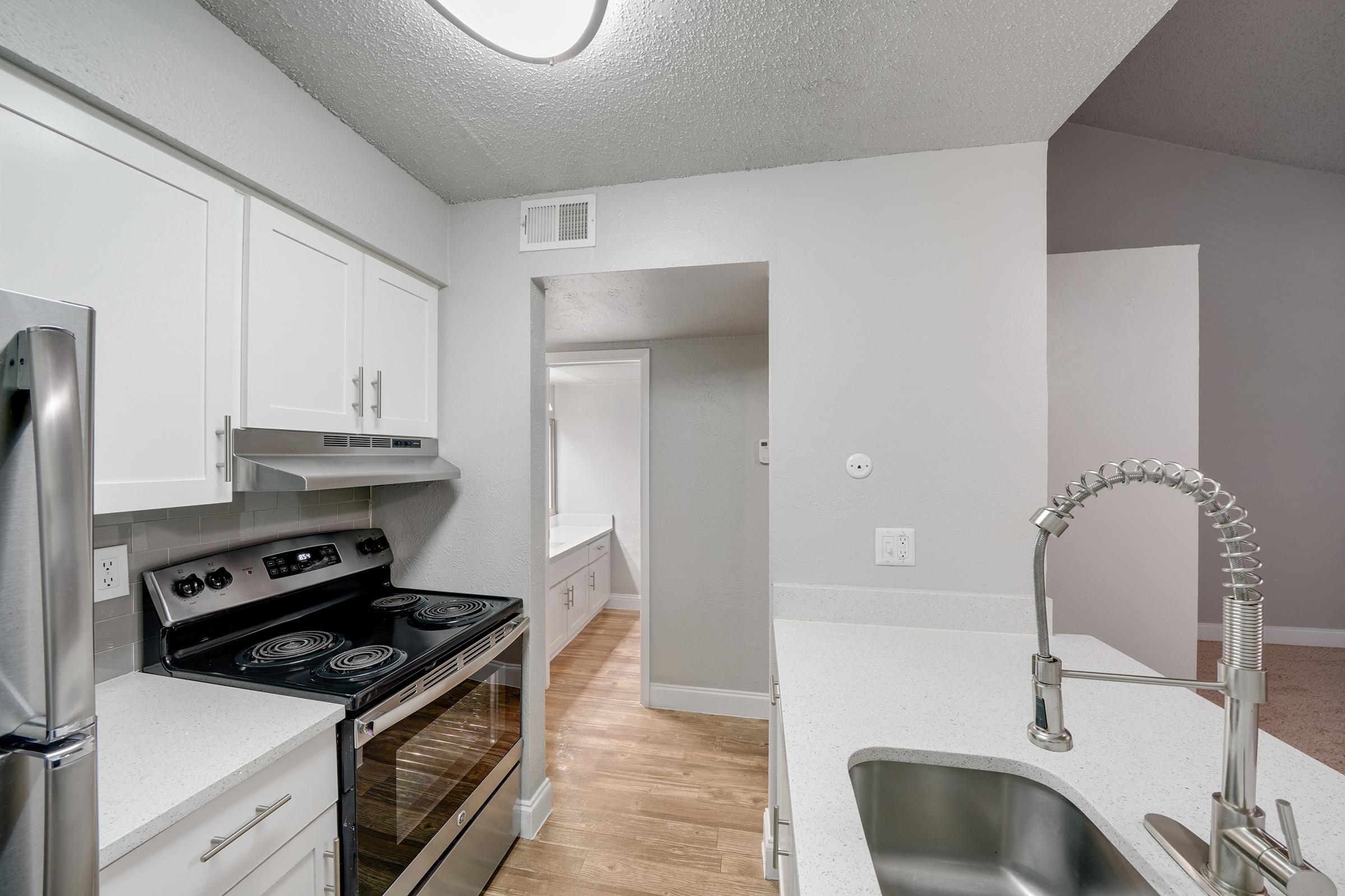 Modern kitchen with white cabinetry, stainless steel appliances including a stove and refrigerator, and a sleek, light countertop. There is a double-basin sink with a modern faucet. The walls are painted in neutral tones, and natural light brightens the space from a nearby doorway.