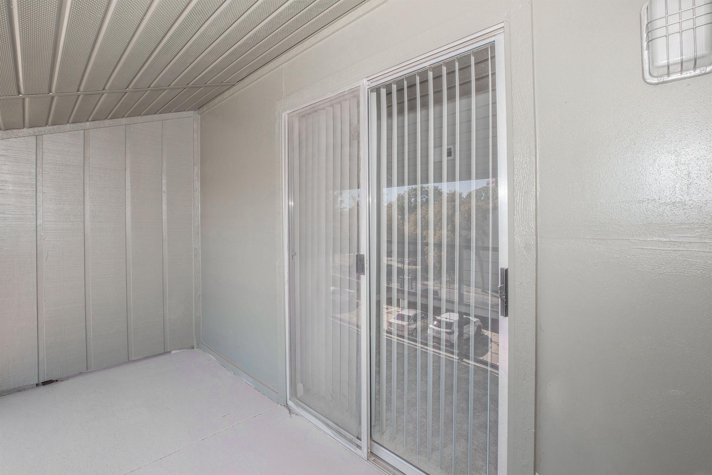 An interior view of a balcony or patio area featuring a sliding glass door with vertical blinds, light gray walls, and a smooth floor. The glass door provides access to an outdoor space, enhancing natural light. The surrounding walls are minimalistic and modern in design.