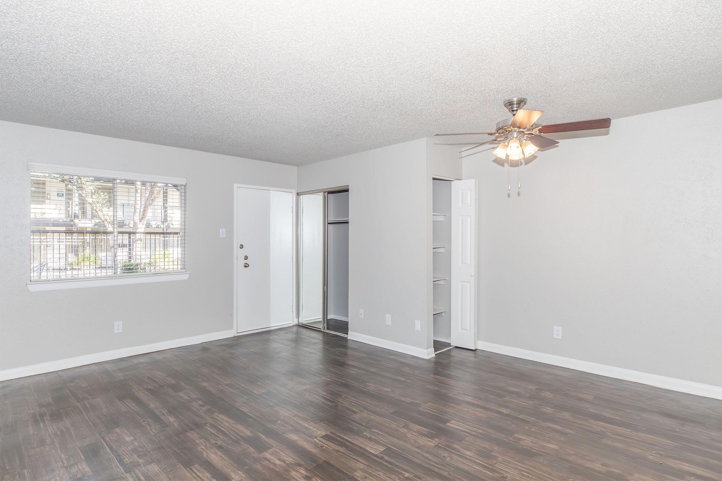 A spacious, well-lit living room featuring a ceiling fan, beige walls, and dark wooden flooring. A window provides natural light, and there is a doorway leading to a closet space. The overall decor is simple and modern.