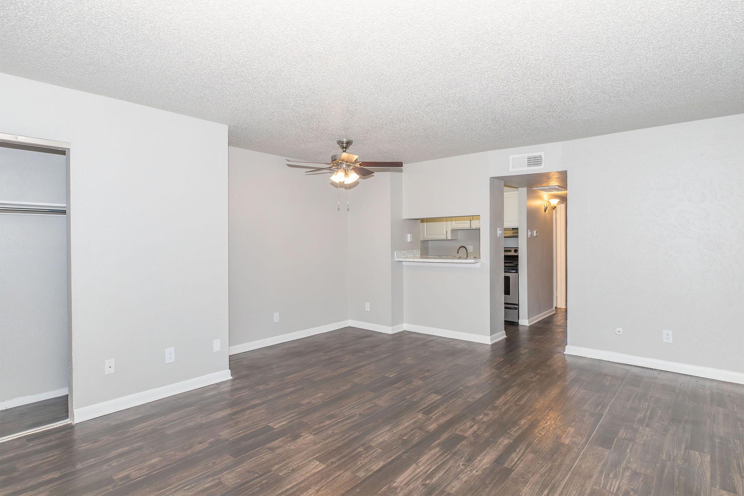 A spacious living room with light-colored walls and dark wooden flooring. A ceiling fan is mounted above, and a bar counter leads to a kitchen area in the background. There is a closet space on the left, enhancing the open and airy feel of the room.