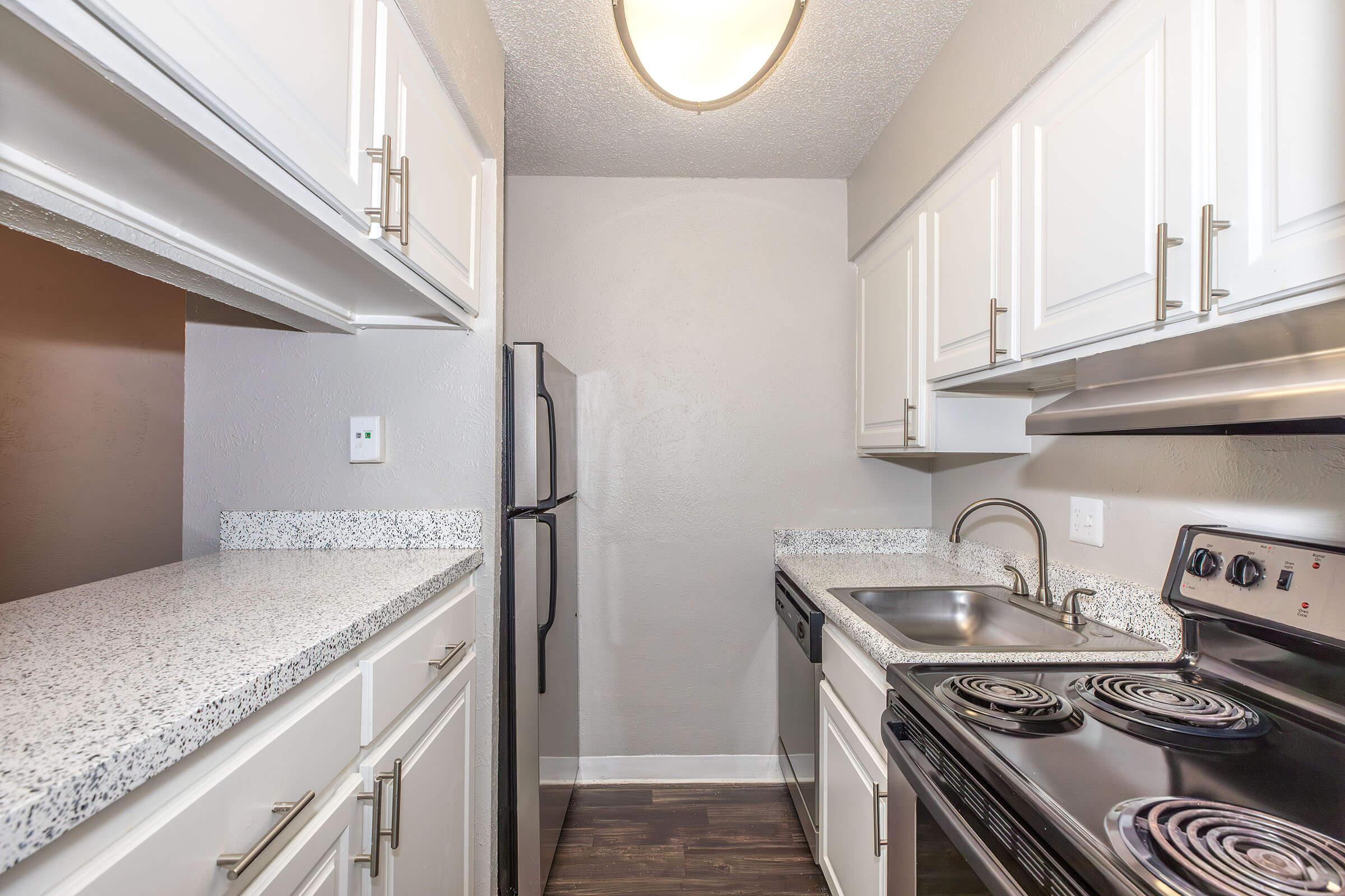 A modern kitchen featuring white cabinets, a granite countertop, and stainless steel appliances. The kitchen includes a black stove, oven, and refrigerator, with a single-basin sink and a light fixture above. The walls are painted in a soft gray tone, creating a bright and inviting atmosphere.