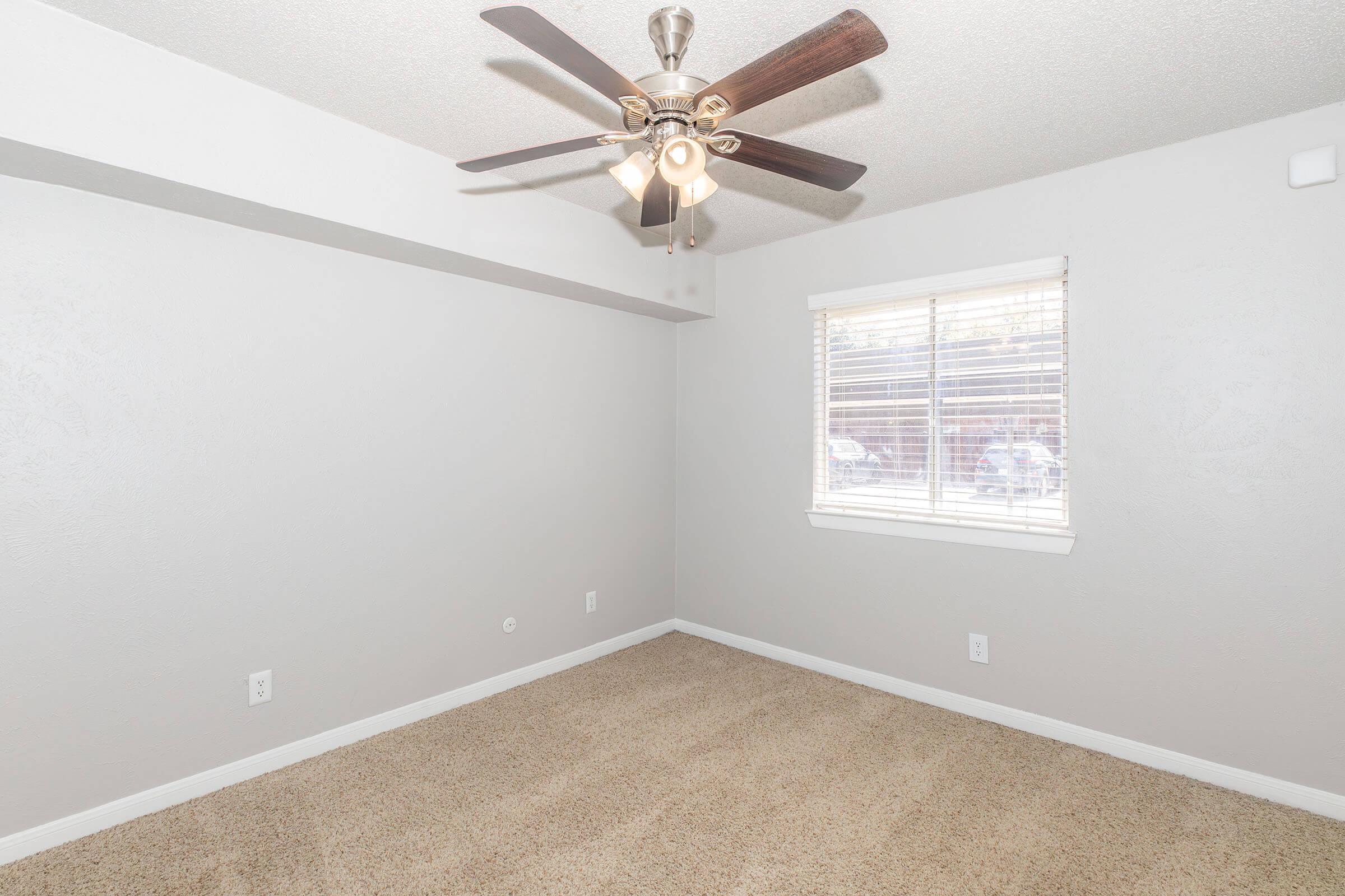 A well-lit, empty room featuring a light ceiling fan and a window with blinds. The walls are painted a light gray, and the floor is covered with beige carpet. The room is spacious with minimal furnishings, providing a blank canvas for decoration or use.