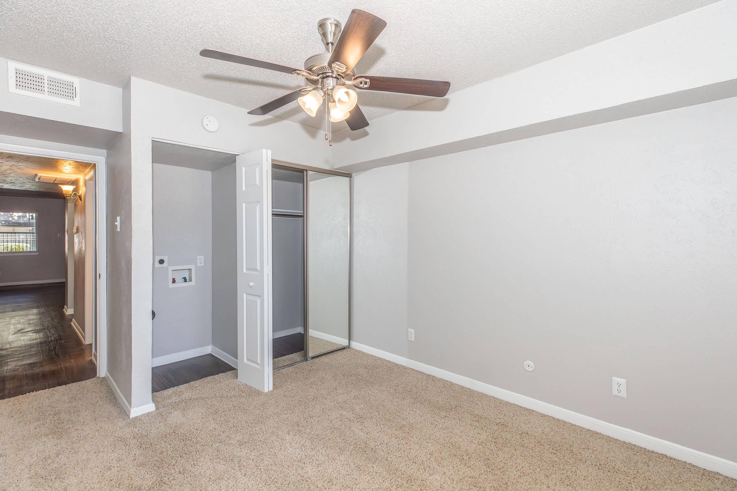 Interior view of a carpeted bedroom featuring a ceiling fan with light, a closet with mirrored doors, and neutral-colored walls. An open doorway leads to another room, and there are light switches and a thermostat visible on the wall. Natural light enters from a window, enhancing the spacious feel.