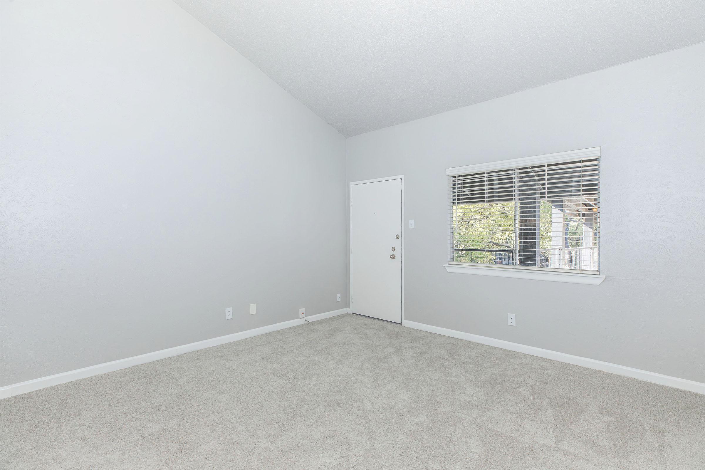 An empty room with light gray walls and carpet. There is a door on one side and a window with horizontal blinds, allowing natural light to enter. The ceiling is high, giving a spacious feel to the room. No furniture or decor is present, emphasizing the bare space.